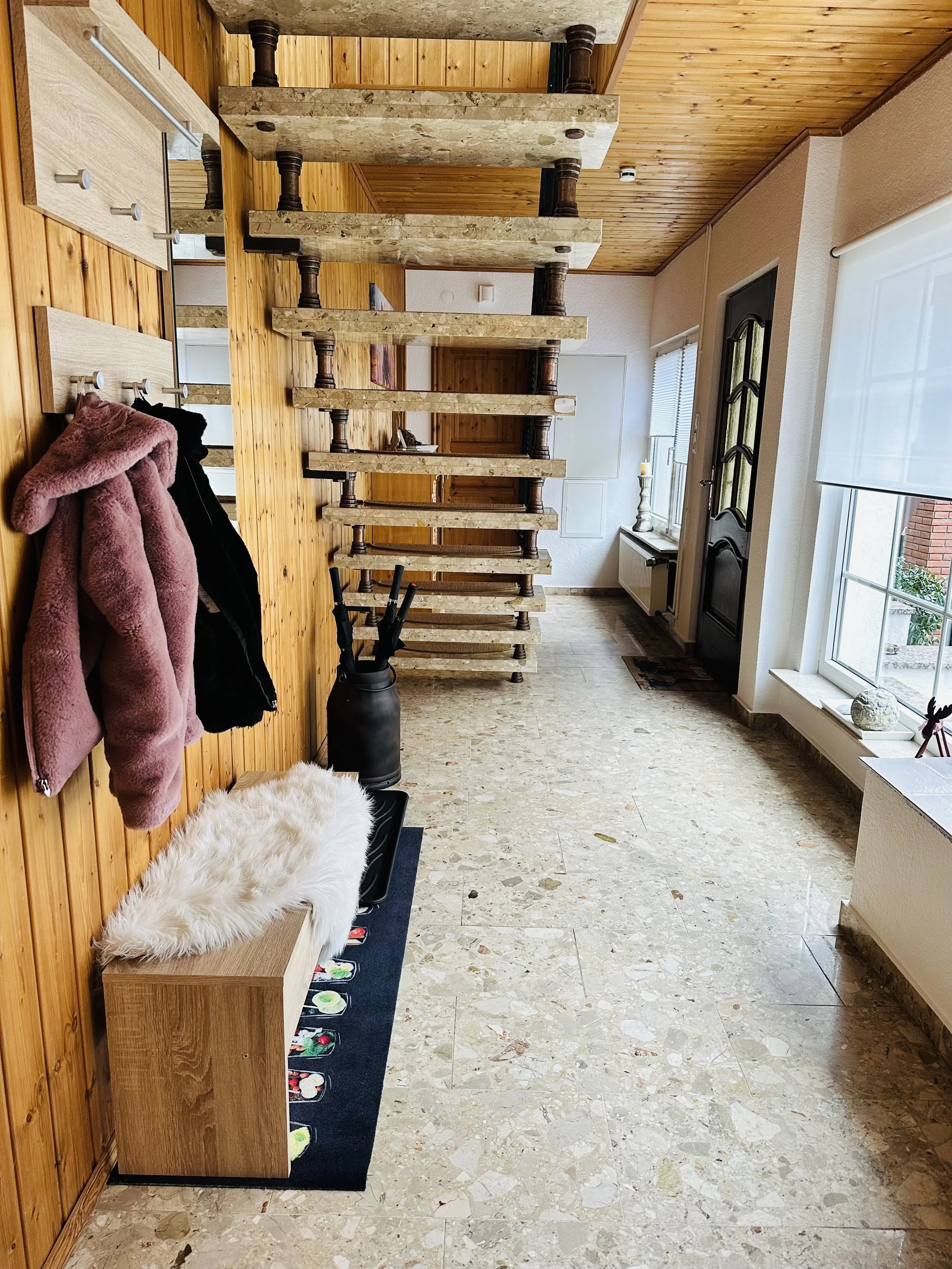Ein Flur mit Holzwand, Marmorboden, einer Garderobe mit Jacken, einem Teppich mit Sushi-Motiv, einem Regal mit Fell, großen Fenstern, einer Haustür und einem Treppenaufgang.