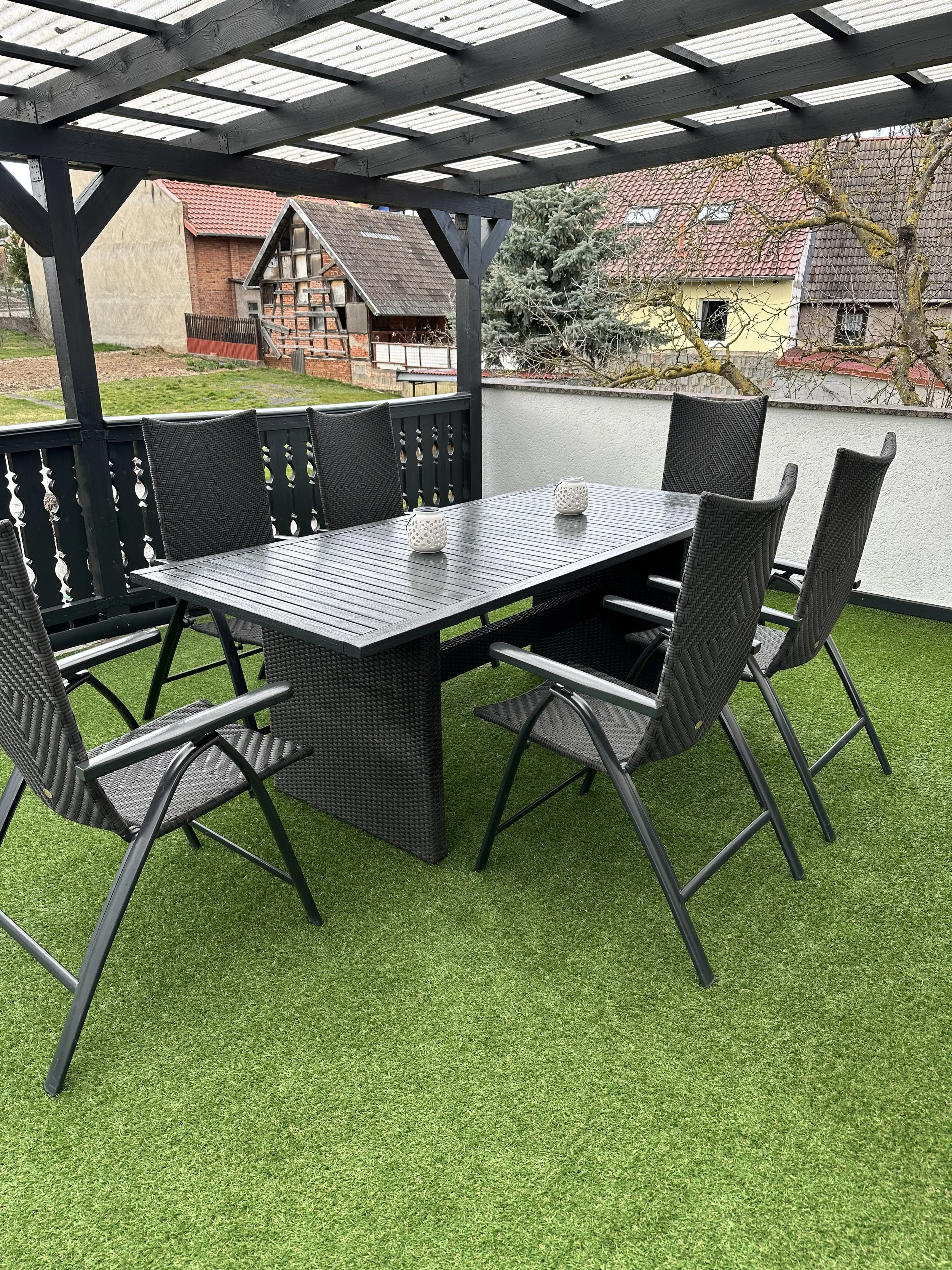 Ein Balkon mit einem schwarzen Gartenmöbel-Set aus einem Tisch und sechs Stühlen auf einem künstlichen grünen Rasen, mit einer weißen Mauern und Bäumen im Hintergrund, darunter eine große Tanne.