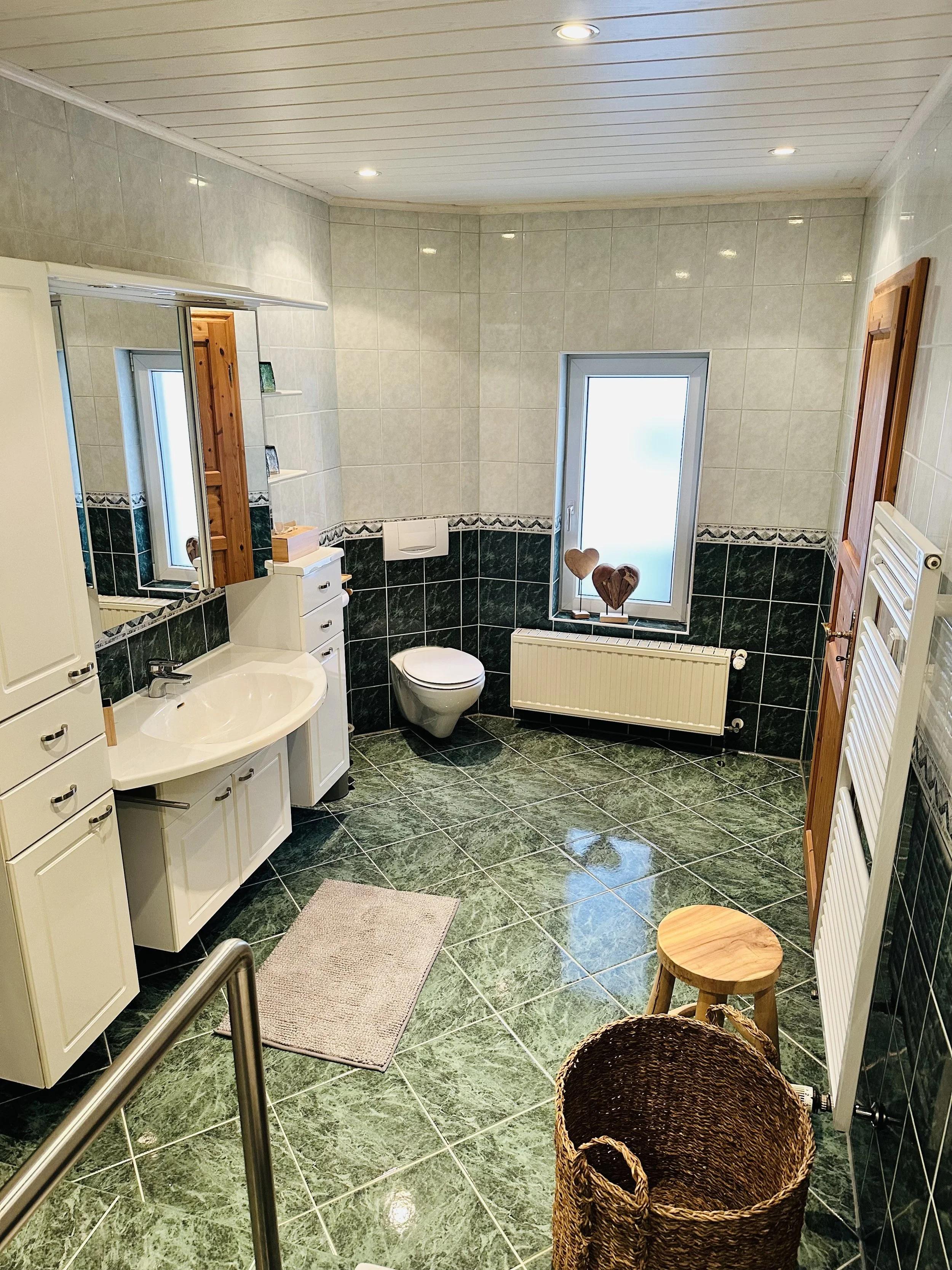 Ein Badezimmer mit weißen und grünen Fliesen, einer weißen Waschtisch mit Unterschrank, einem weißen WC, einem großen Fenster mit Fensterbank, einem Wandradiator, einem Spiegel, einem kleinen Stehpult, einem Korb und einem Hocker