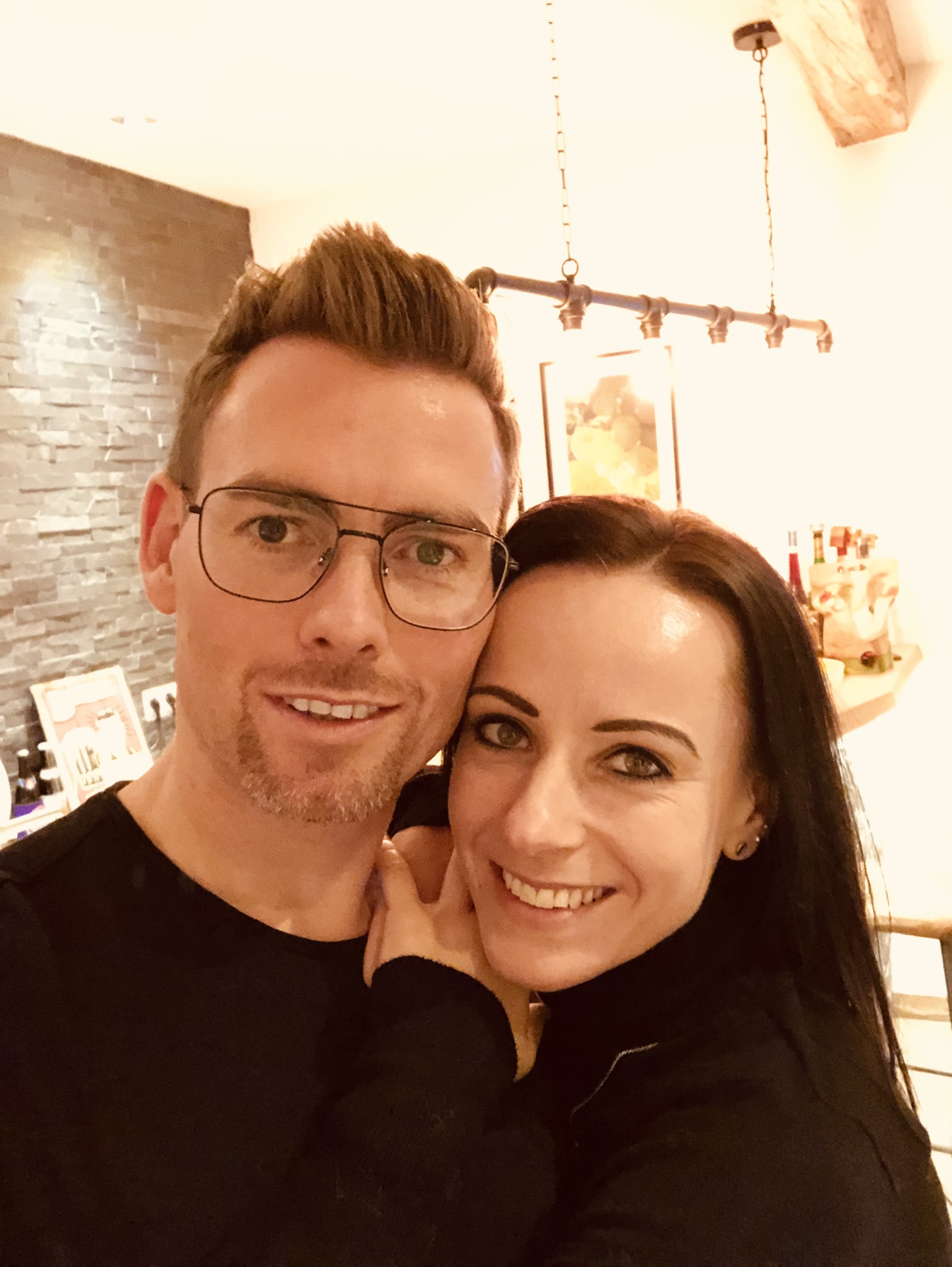 Ein Mann mit Brille und ein Frau mit dunklen Haaren posieren lächelnd für ein Selfie in einem liebevoll dekorierten Raum.