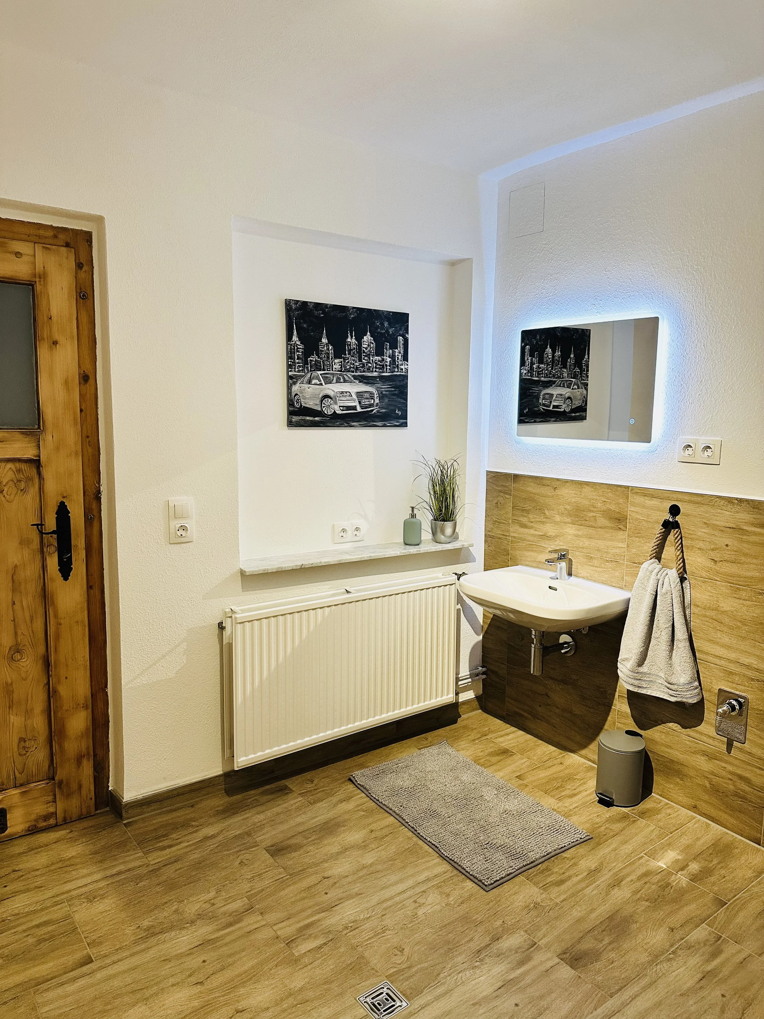Ein Badezimmer mit Holzfußboden, einem weißen Waschbecken, einem Spiegel mit LED-Beleuchtung, Wandbilder mit schwarzen und weißen Stadtansichten, einem Handtuch an einer Halterung, einem Teppich, einer Heizung, einem Wäschekorb und einer kleinen Pfla