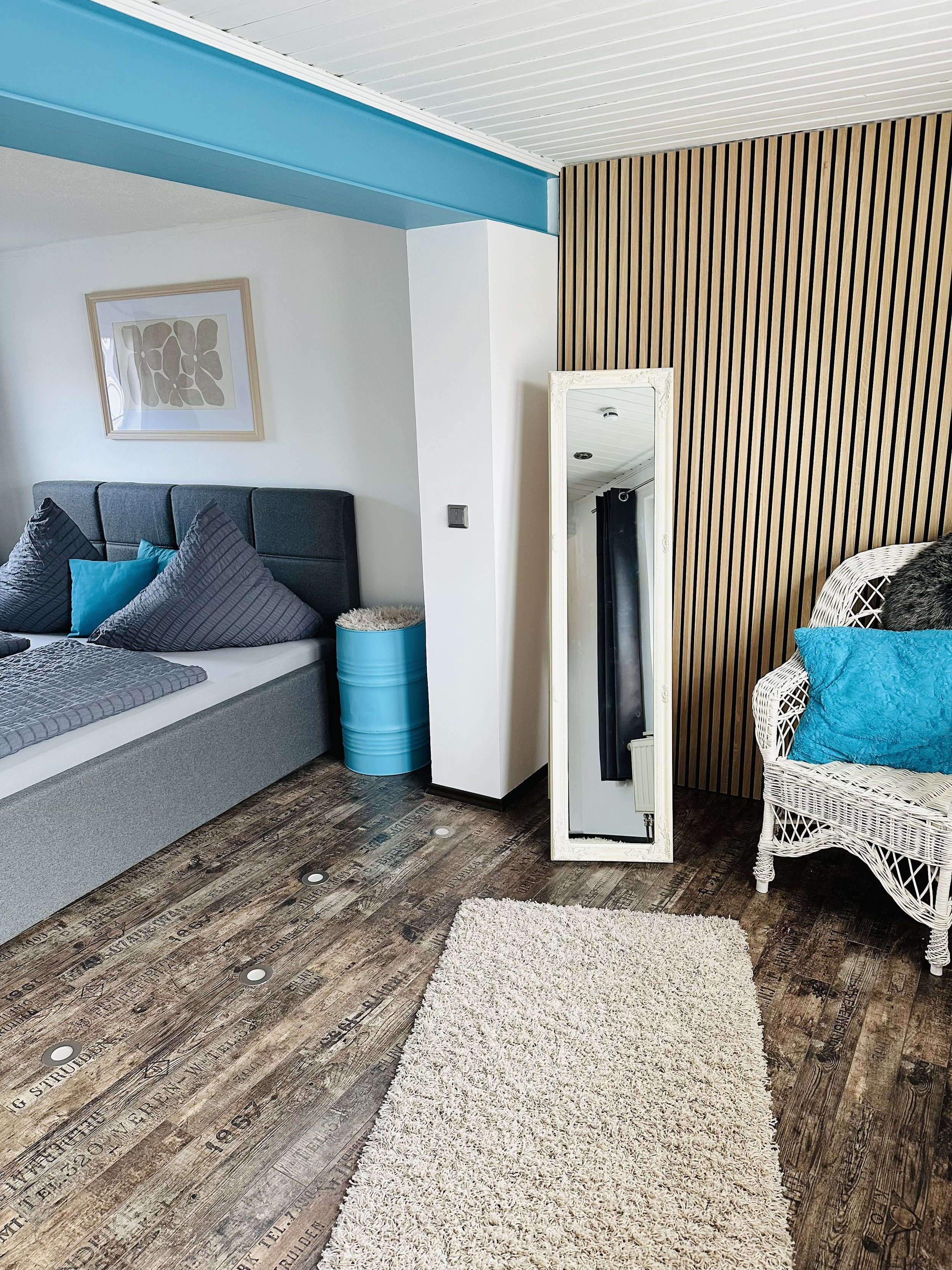 Ansicht eines Wohnzimmers mit Holzfußboden, einem weißen Teppich, einem schwarzen Bett mit Kissen und Polster, einer weißen Korbwäsche, einem weißen Spiegel, Holzverkleidung an der Wand und einem weißen Wandrahmen mit Kunstwerk.