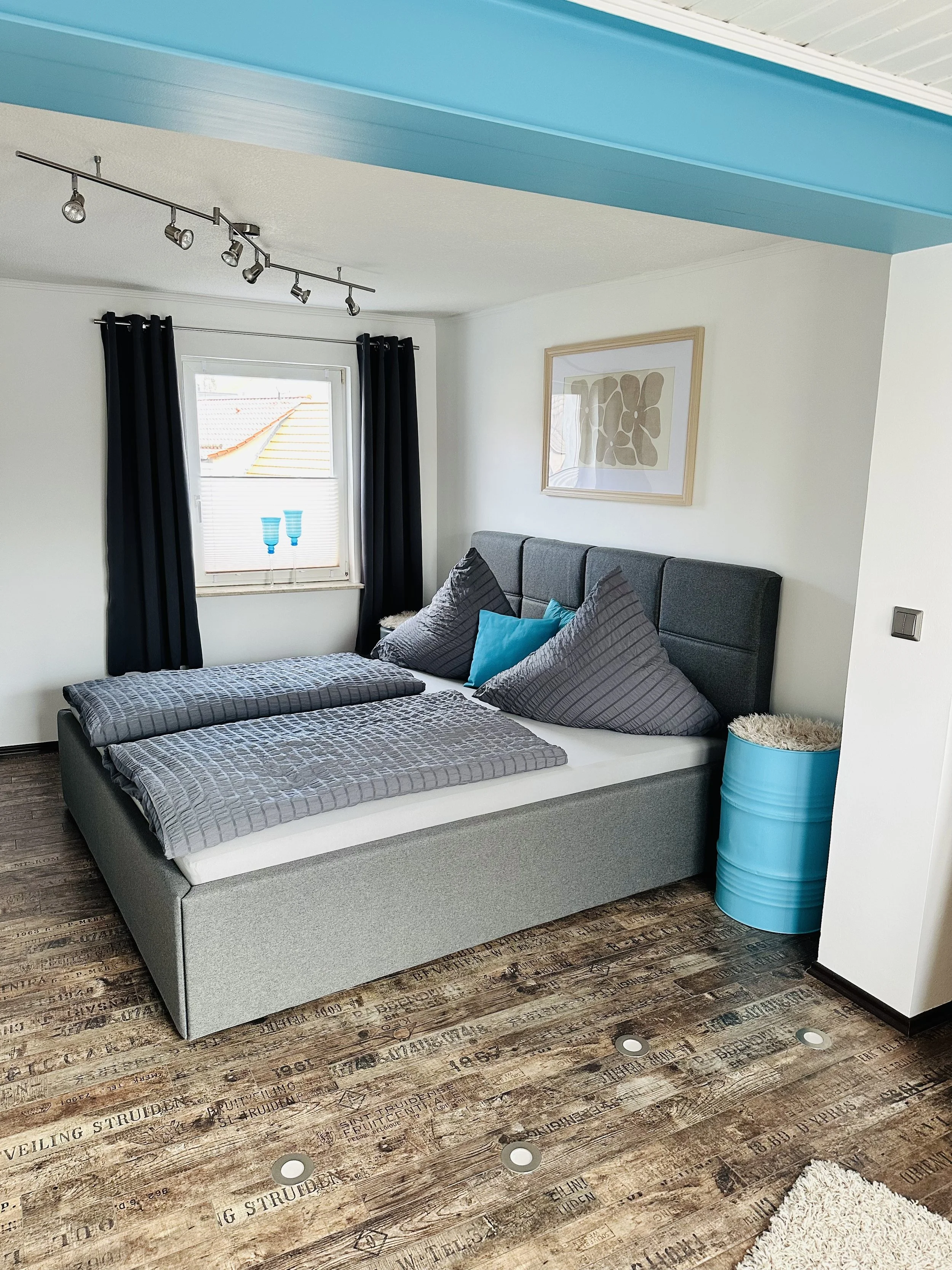 Ein Schlafzimmer mit einem Doppelbett, schwarzen Vorhängen, einer Wandkunst, einem blauen Metalleimer und einem Fenster mit blau-weißen Vasen. Das Zimmer hat Holzboden mit Aufdrucken und Bodenleuchten.