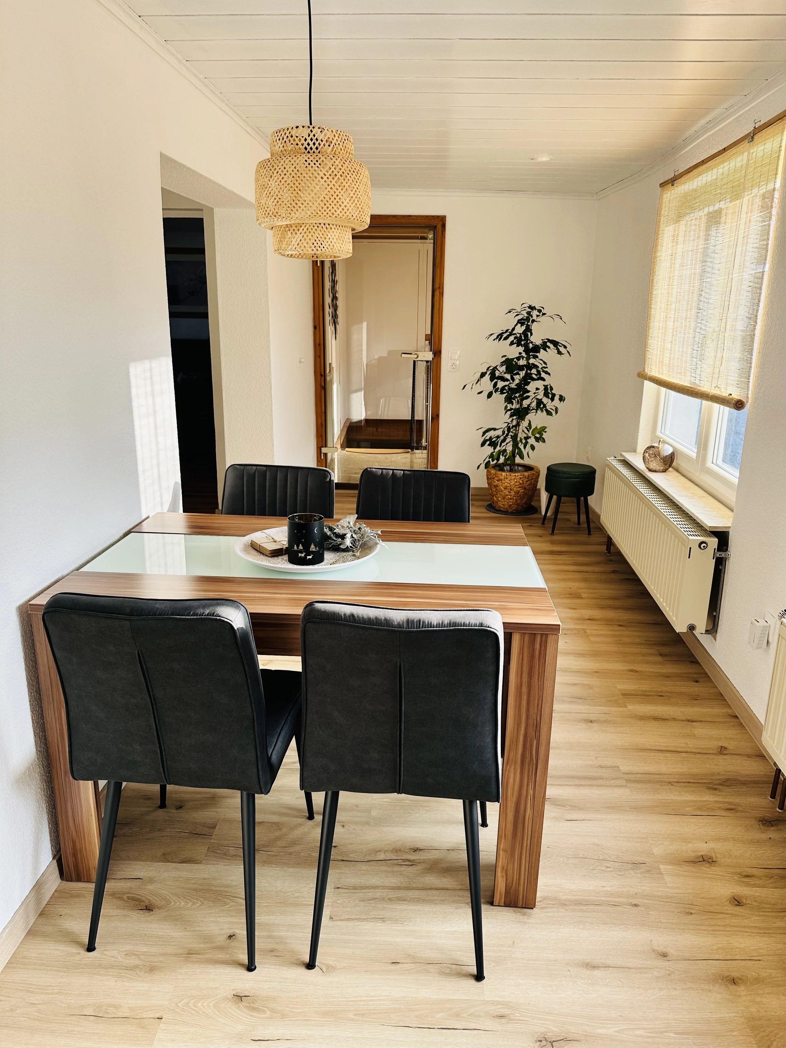 Ein Esszimmertisch mit schwarzen Stühlen, eine Topfpflanze, eine Fensterbrüstung mit Dekogegenständen, ein Leuchter, eine Tür im Hintergrund, und Holzfußboden.