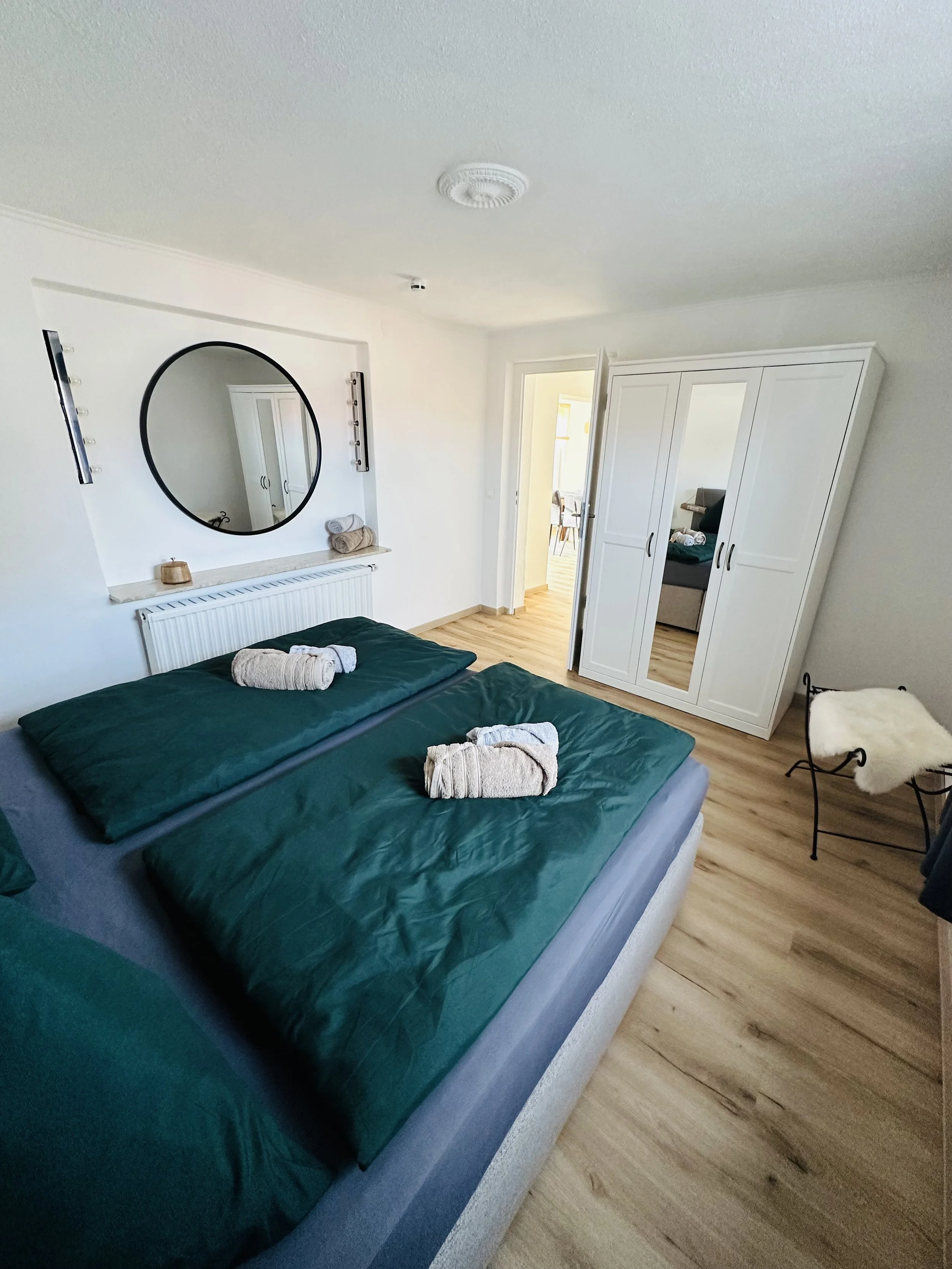 Ein Schlafzimmer mit einem Doppelbett, grüner Bettwäsche, Handtüchern und einem Spiegel an der Wand. Es gibt einen weißen Kleiderschrank und eine kleine Sitzgelegenheit.