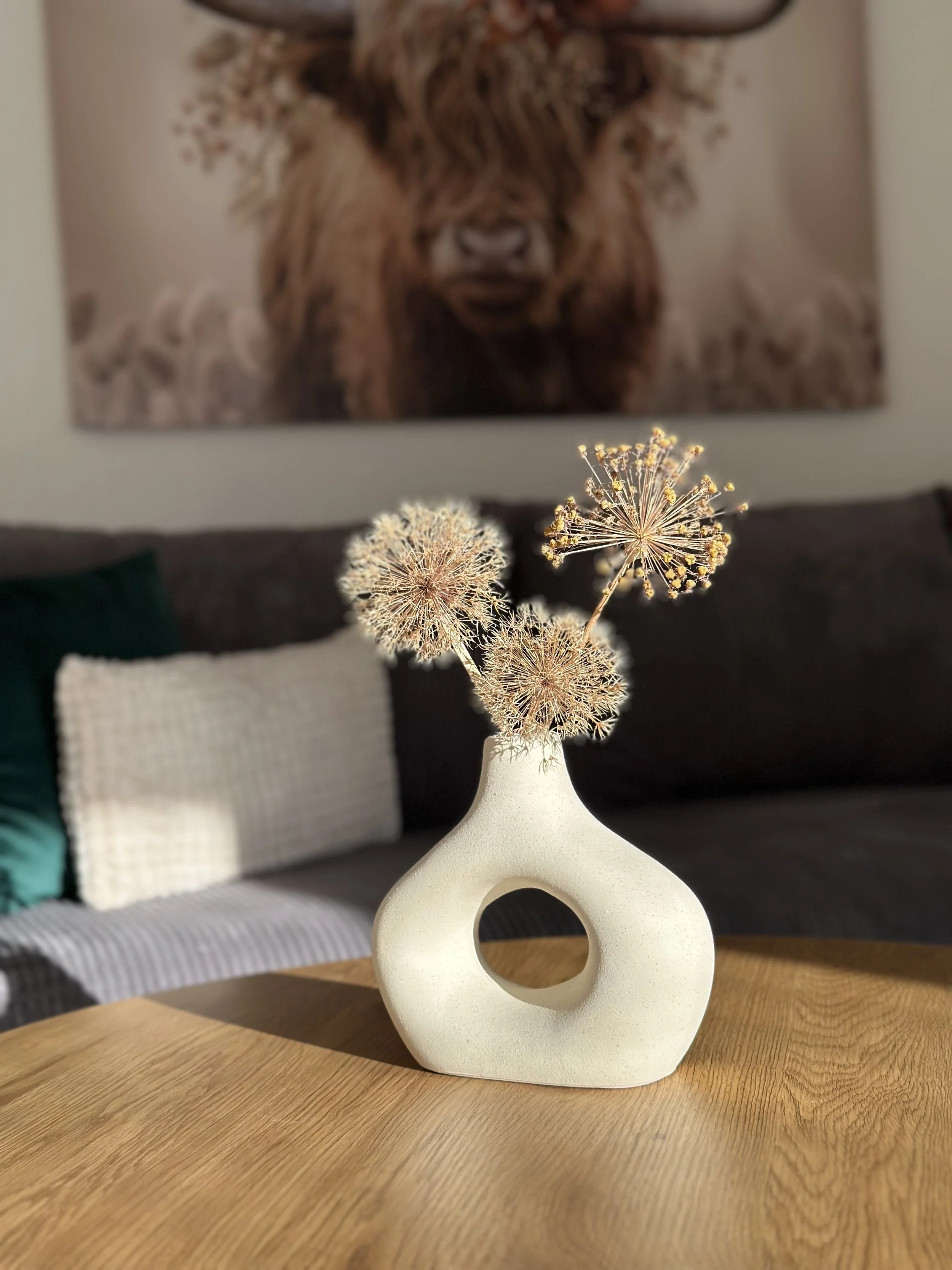 Weiße Vase mit geometrischer Form auf Holzoberfläche, enthält Verblühte Gartenblumen, im Hintergrund ein Tryptichon mit Bärenbild auf Wand.