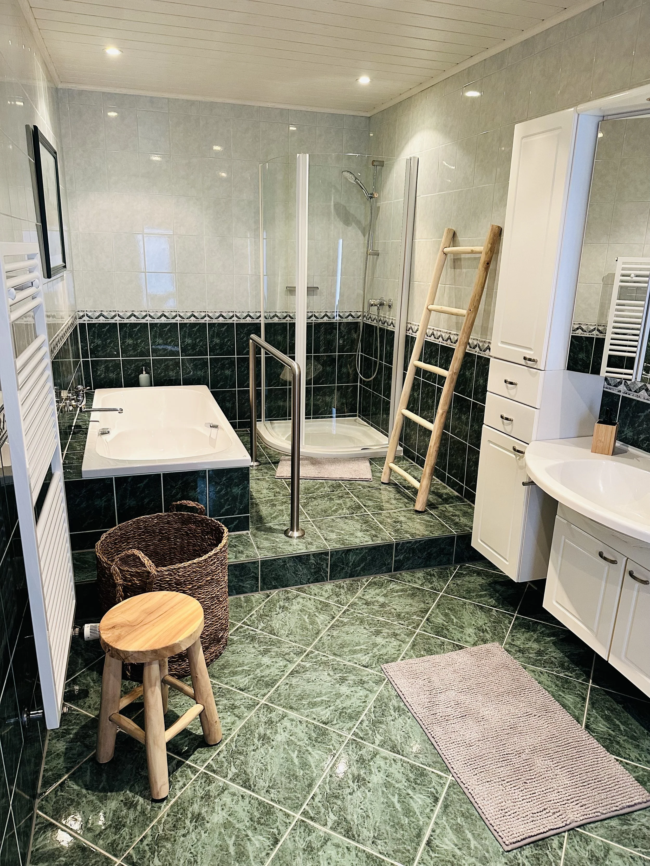 Ein Badezimmer mit grünen Marmorfliesen, Badewanne, Dusche, weißem Waschtisch, Leiter, Holzhocker, Körbe, Handtücher, Spiegel und Schränken.