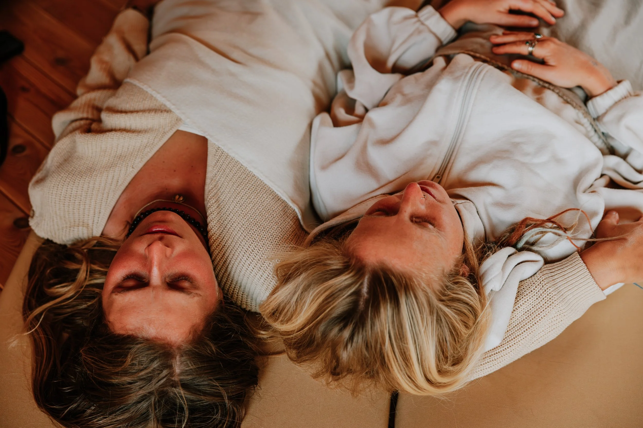 Four women lie close together on a tan surface, with their heads touching, their eyes closed, appearing relaxed and content.