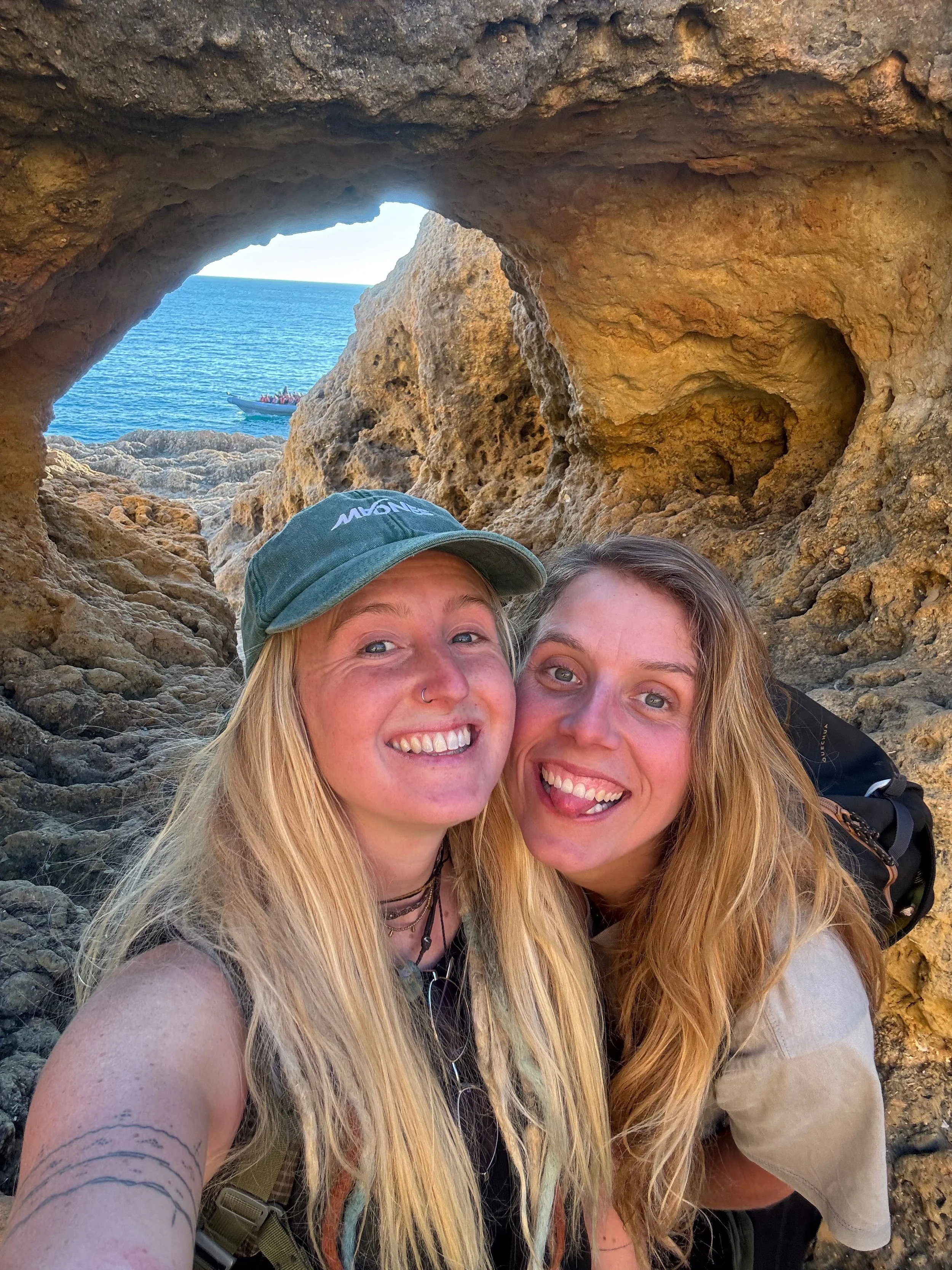 Sanne en Charlene van Sisterhood Marketing nemen een selfie tussen rotsen met uitzicht op de zee, een boot met mensen vaart op de achtergrond.