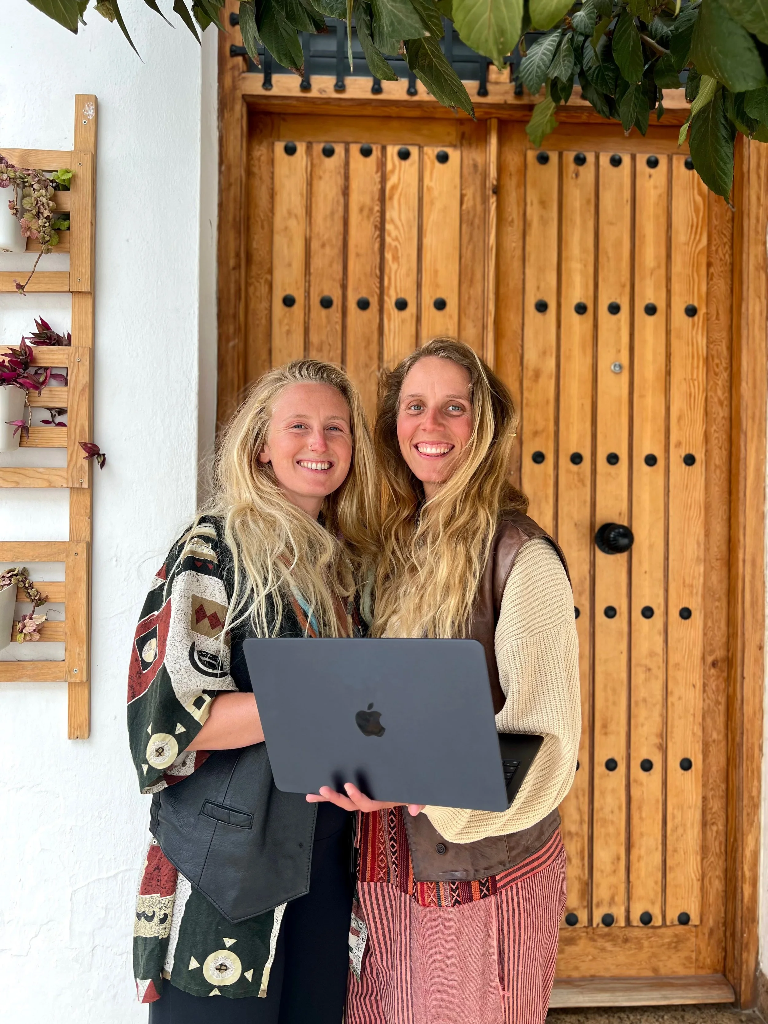 Two women standing together outside near a wooden door and a white wall with wall-mounted planters, holding a laptop and smiling at the camera.