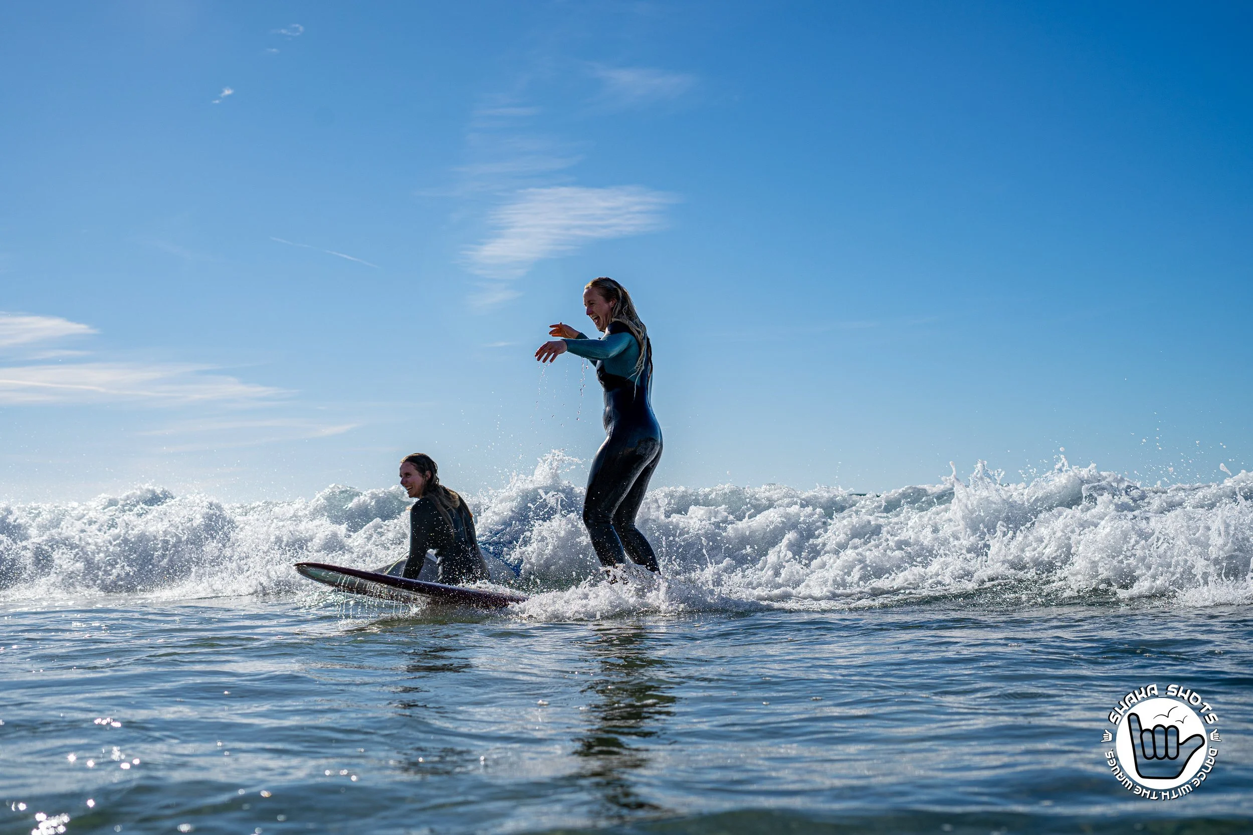 Sanne en Charlene van Sisterhood Marketing genieten van het surfen op de oceaan, met een helderblauwe lucht op de achtergrond.