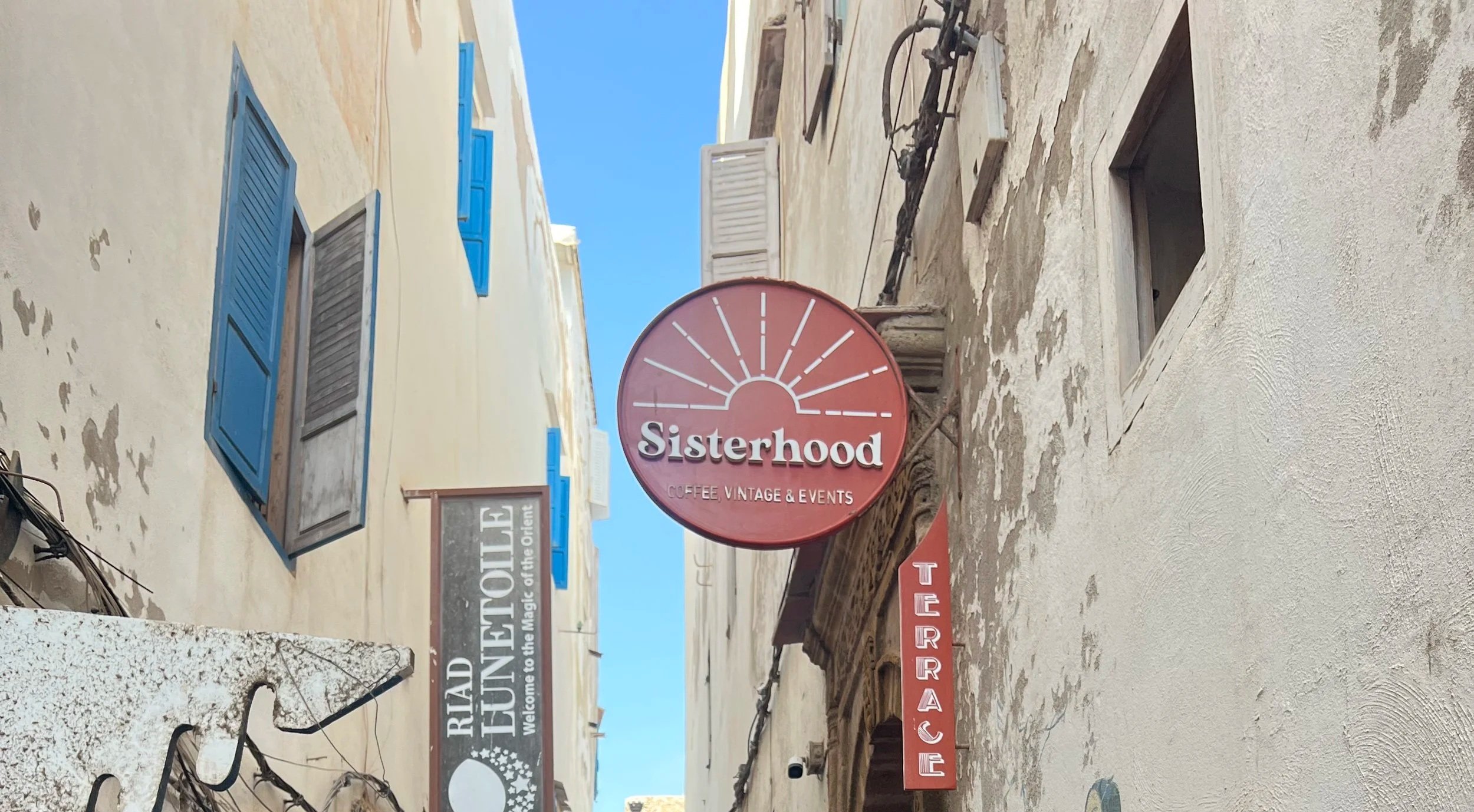 A street scene with a pink circular sign reading 'Sisterhood Coffee Vintage & Events', a beige building with blue window shutters, and other signs including 'Welcome to the Magic of the Ocean' and 'Gourmet'.