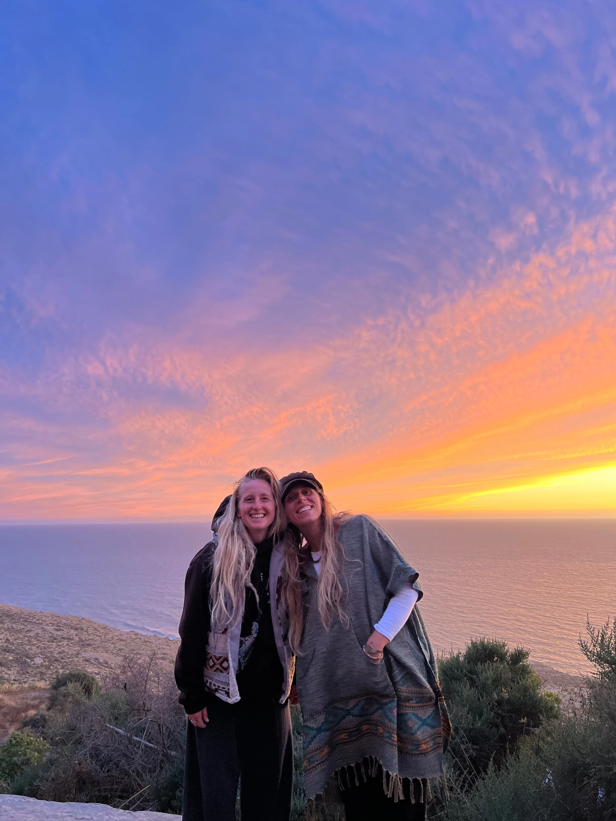 Two women standing close together outdoors at sunset near a body of water with a colorful sky featuring pink, purple, and orange hues. They are smiling at the camera, with one woman wearing a black jacket and the other in a grey poncho and patterned skirt, and they have long hair.