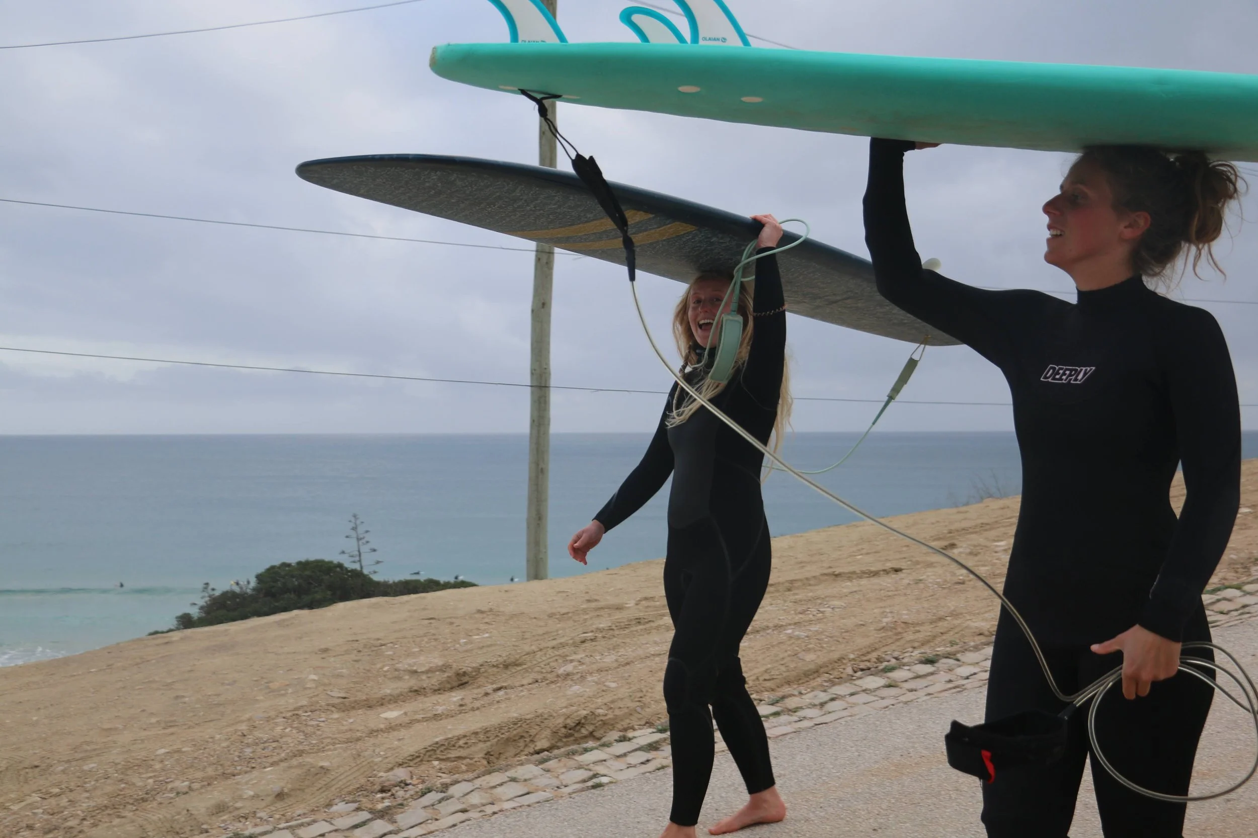 Twee vrouwen lopen langs de kust, beiden dragen wetsuits en dragen surfplanken op hun hoofd.