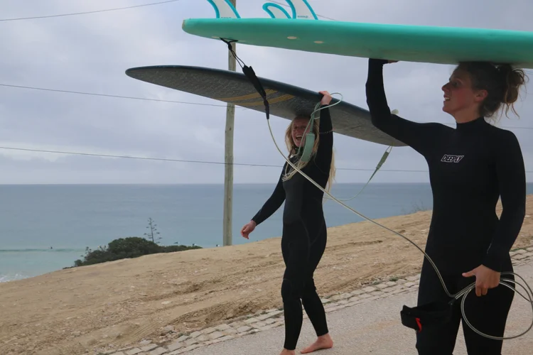 Sanne en Charlene lopen in wetsuits langs de kust met surfplanken op hun hoofd