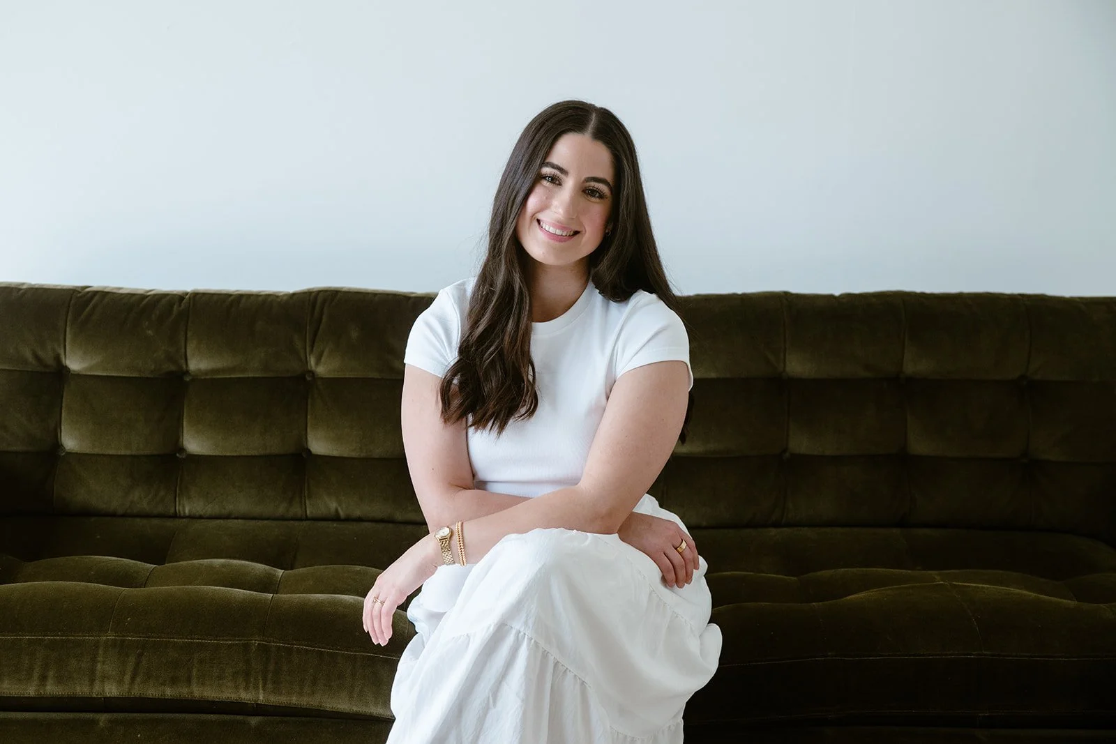 A young woman with long dark hair, wearing a white t-shirt and white skirt, sitting on a vintage green velvet sofa, smiling, with a plain light-colored wall in the background.