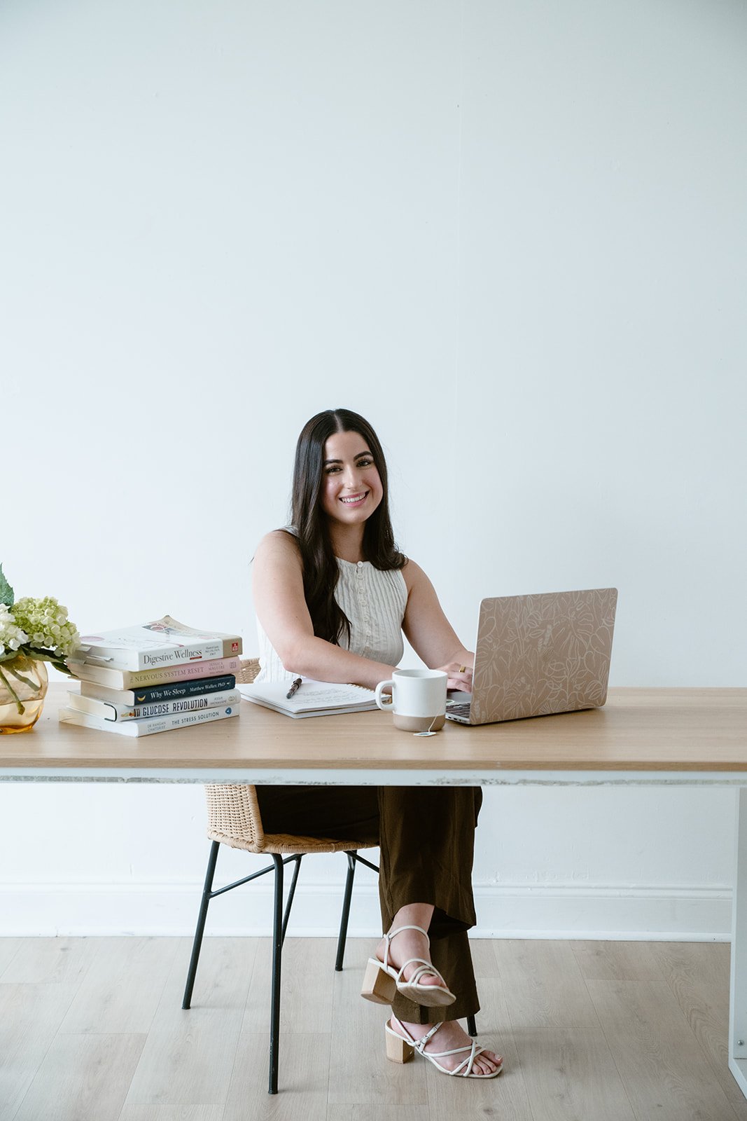 A woman with long dark hair, smiling, sitting at a desk with a laptop, a white mug, and a stack of books, in a minimalist room.