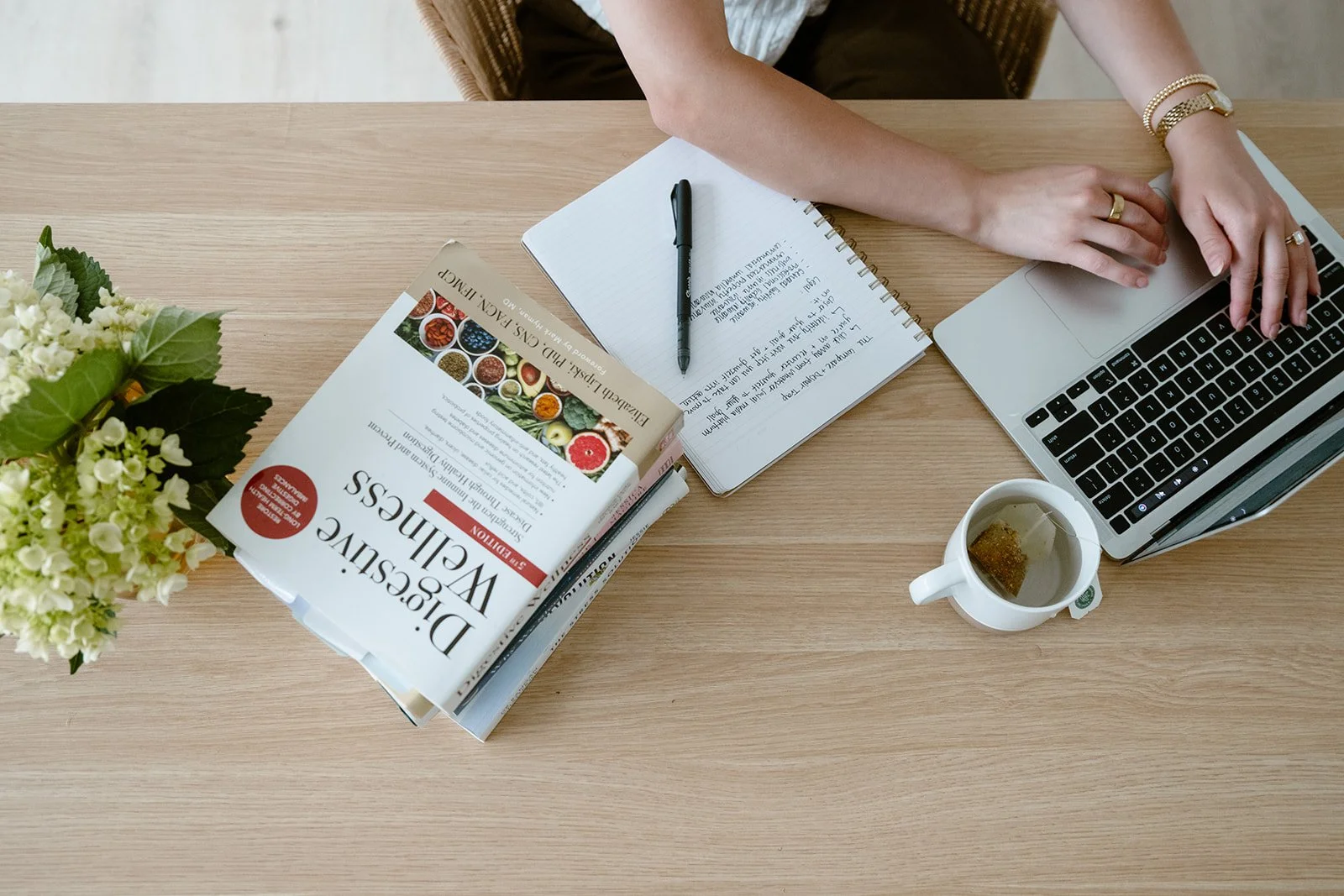 Top view of a wooden desk with a person working on a laptop. The desk has a plant, a coffee mug with tea, a notebook with handwritten notes, a black pen, and a book titled 'Diseases of Vegetables'.