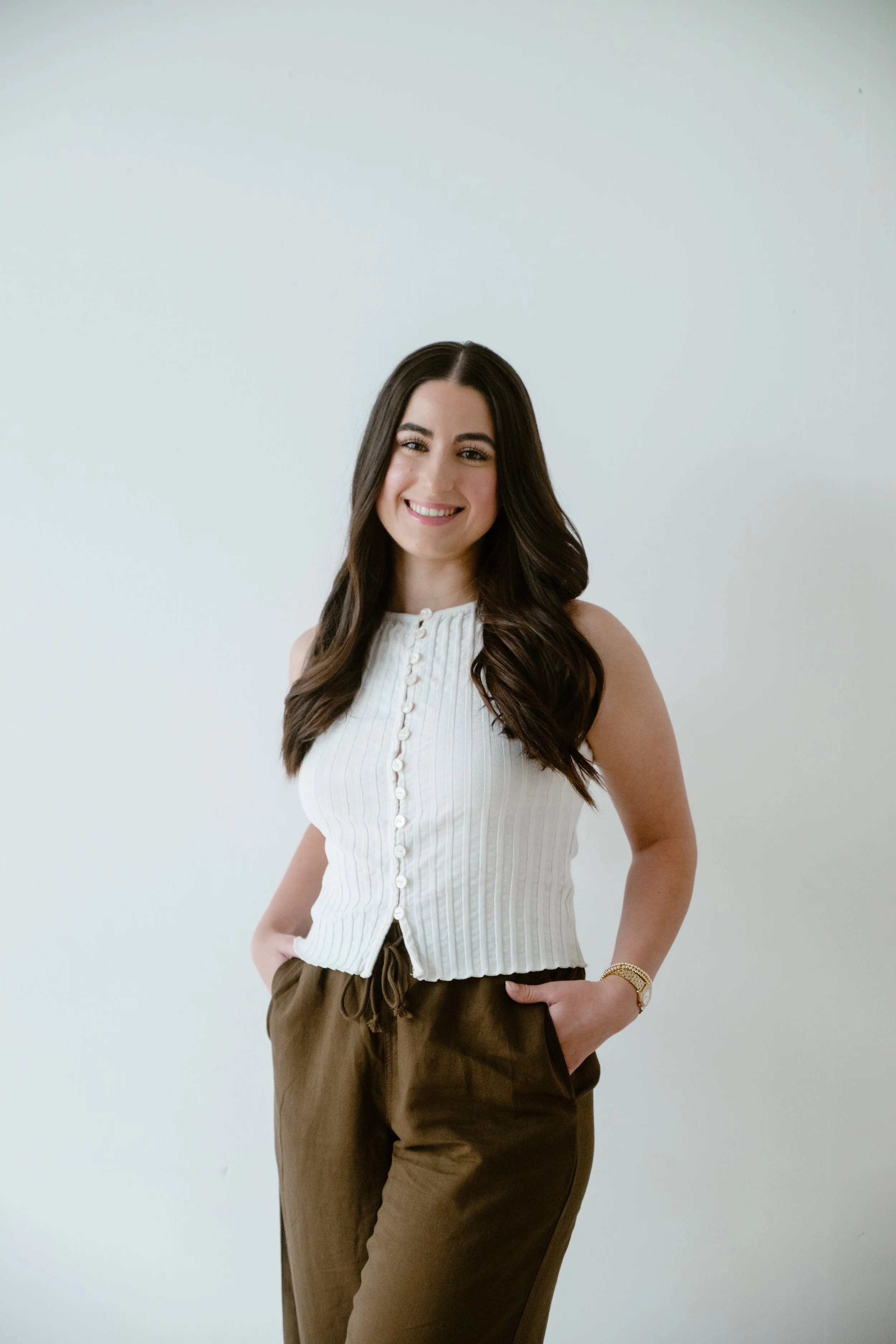 A woman smiling and standing with her hands partially in her pockets against a plain white wall.