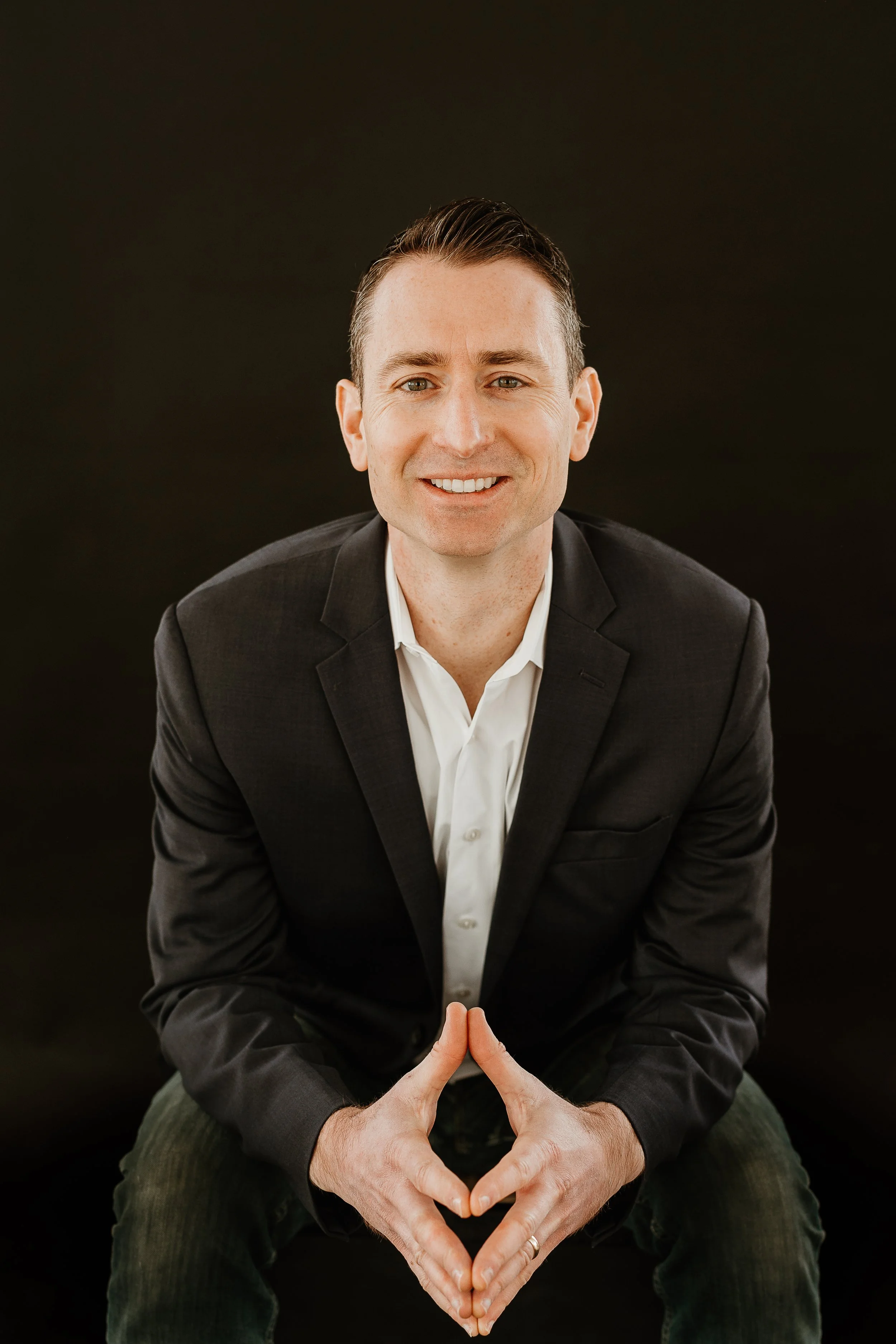 A man in a suit sitting on a chair against a black background, smiling, with his hands forming a heart shape in front of him.