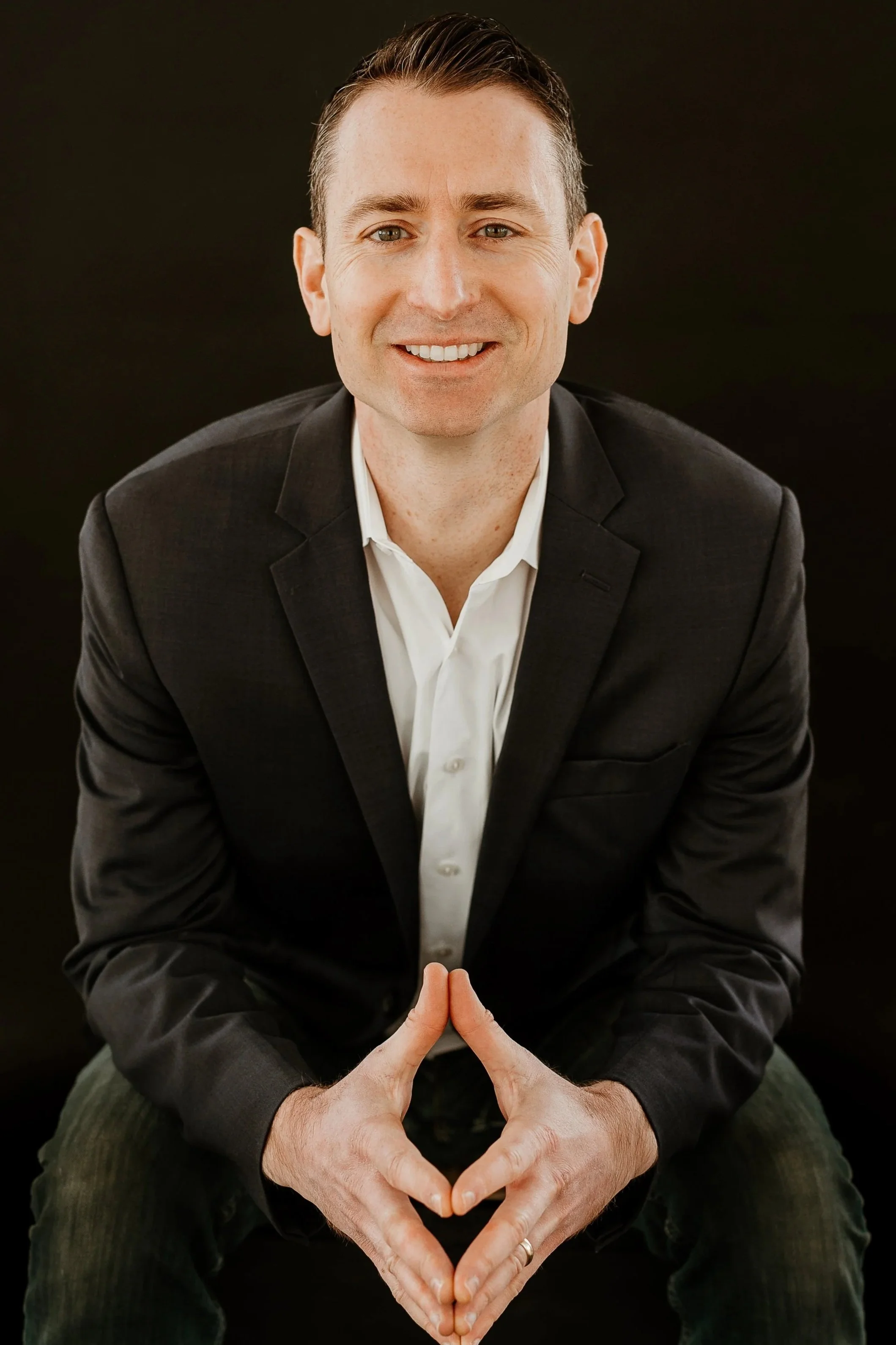 A smiling man with short dark hair, wearing a black suit jacket and white shirt, sitting against a black background. His hands are positioned in front of him, forming a heart shape with his fingers.