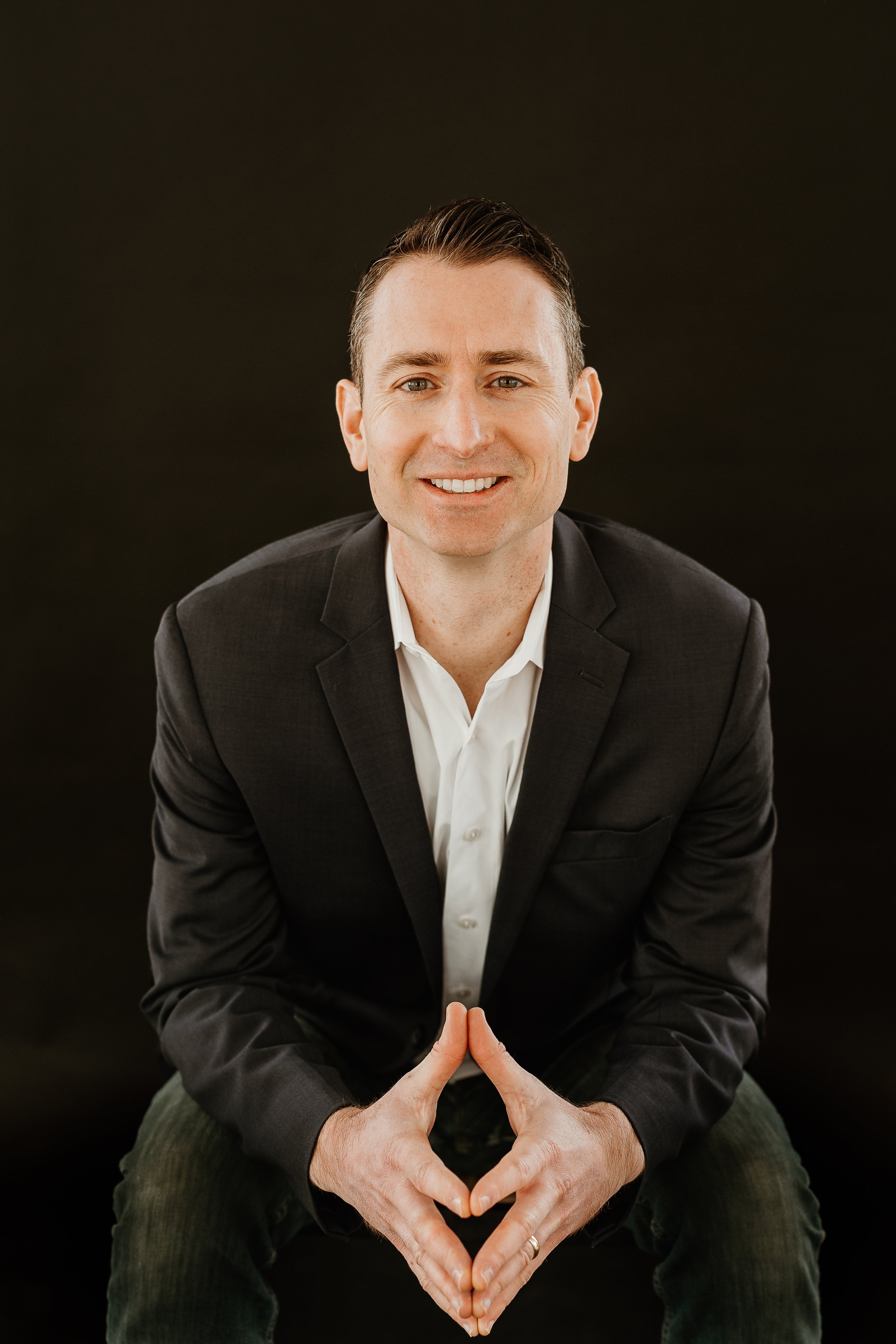 A man in a black suit and white shirt is smiling, with his hands forming a heart shape. He is seated against a black background.