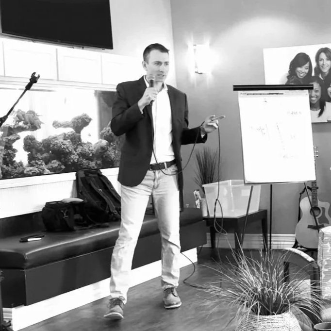 Man in blazer and jeans giving a presentation with a microphone and smartphone, standing beside a whiteboard in a room with a guitar, a fish tank, and photos on the wall.