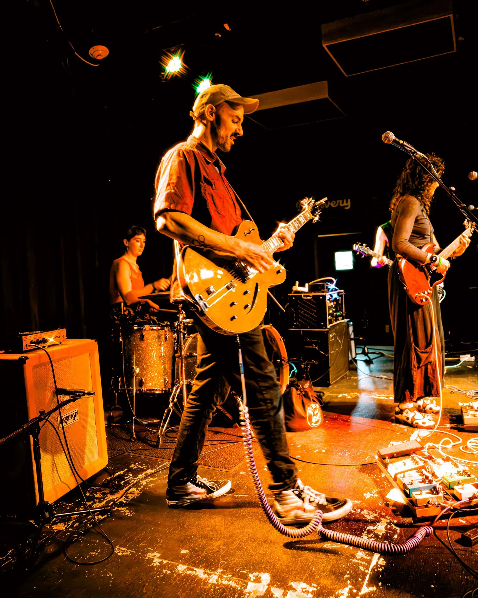 Musicians performing on stage with guitars, keyboard, and drums, surrounded by amplifiers and equipment, under dim lighting with colorful stage lights.