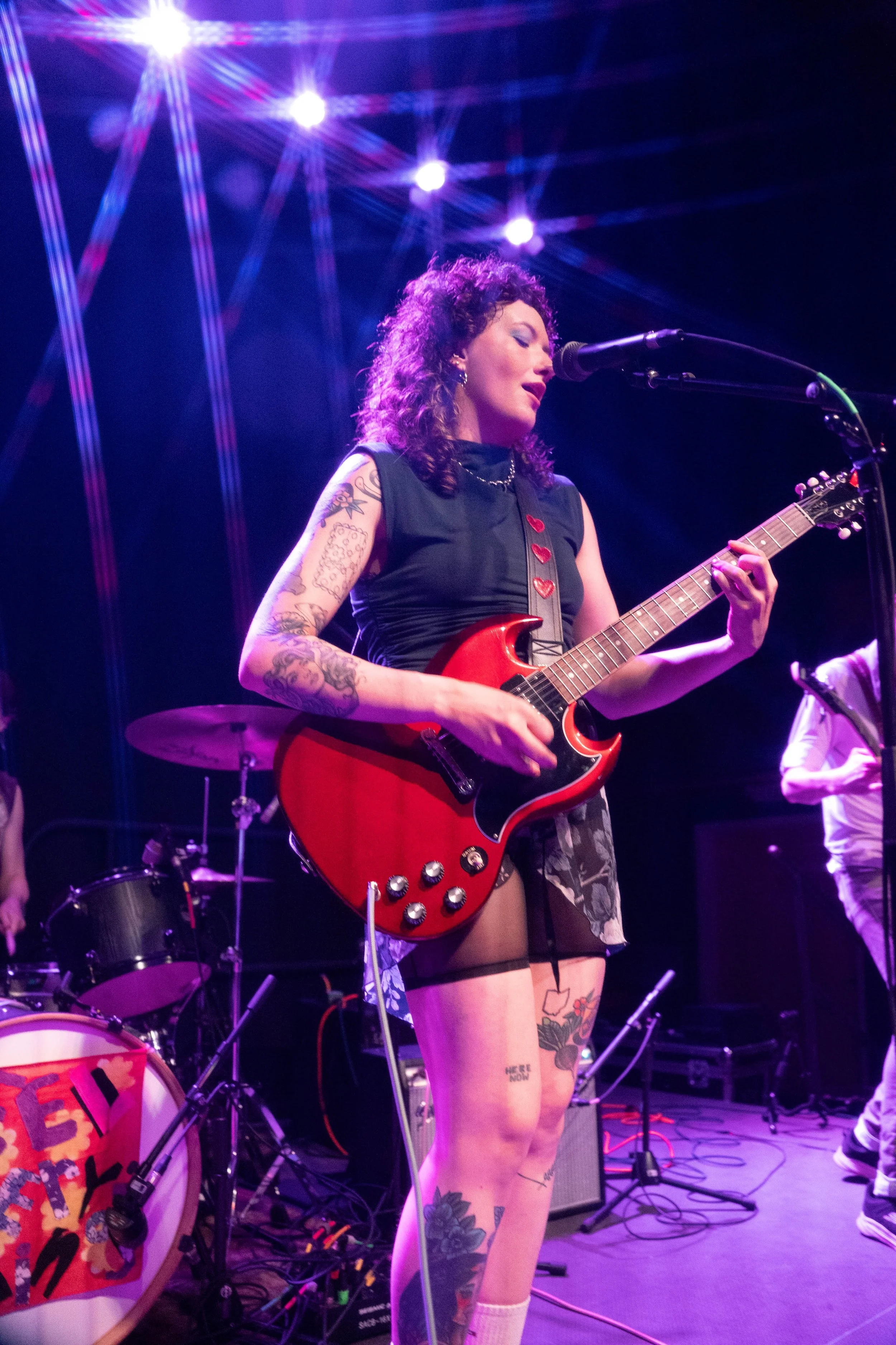 A woman with tattoos playing an electric guitar on stage under purple stage lights, wearing a sleeveless black top and a short skirt.
