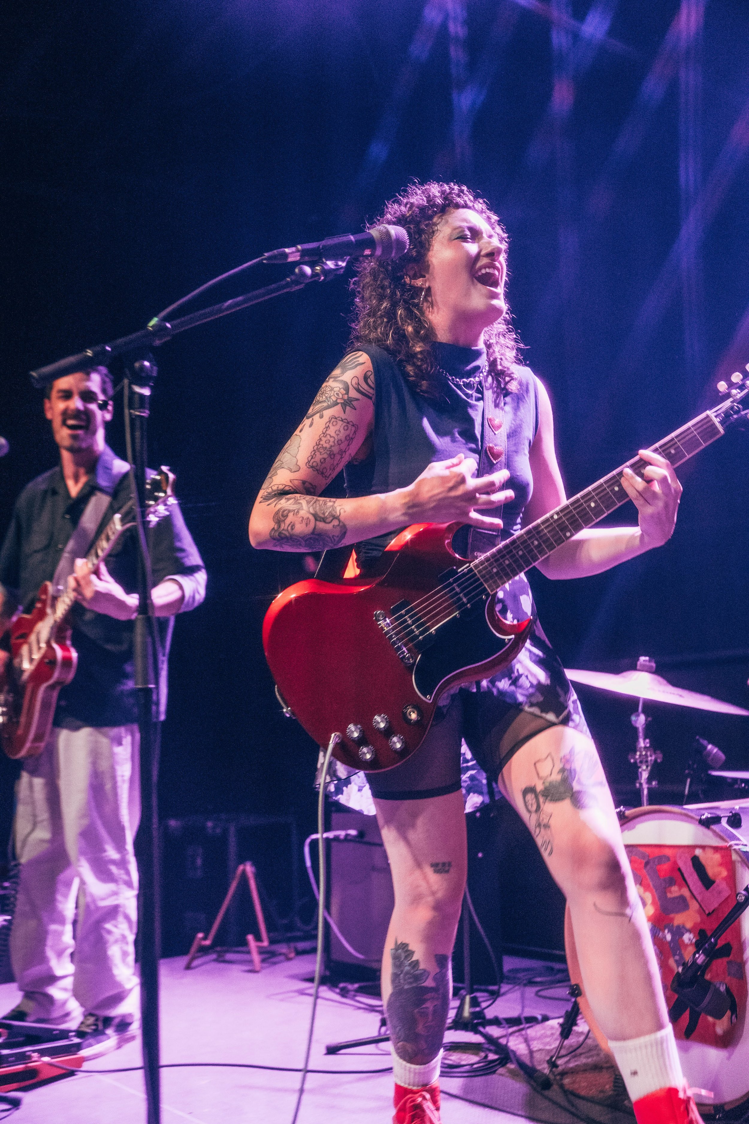 A woman with curly hair, tattoos, and wearing a sleeveless top passionately singing and playing red electric guitar on stage. A man in the background also plays guitar, dressed in a dark shirt and light-colored pants. Stage lights cast a purple hue.