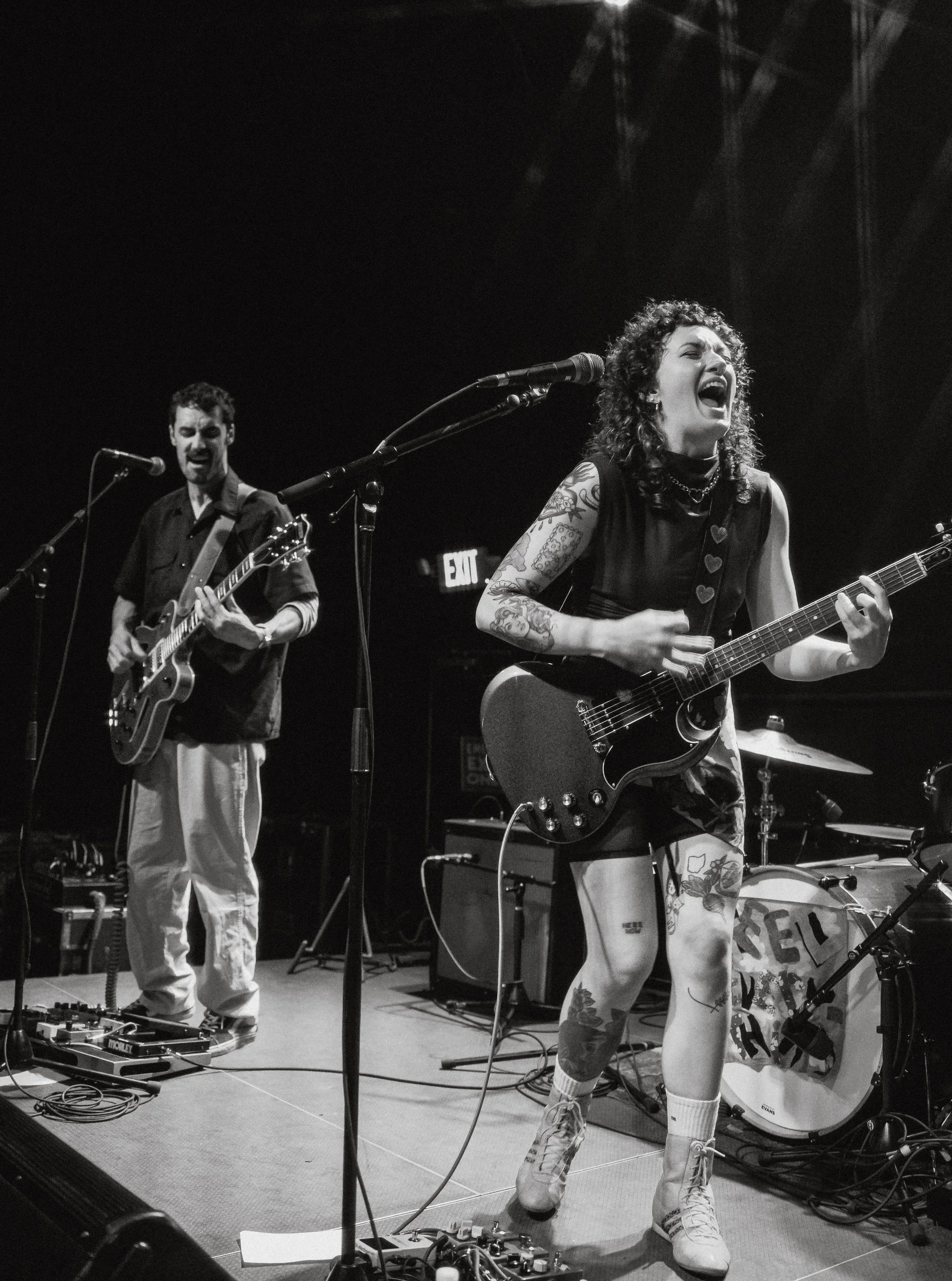 A female musician passionately singing and playing an electric guitar on stage, with tattoos on her arms and legs, and wearing sneakers. She is accompanied by a male guitarist in the background, both performing under stage lights with musical equipment around them.