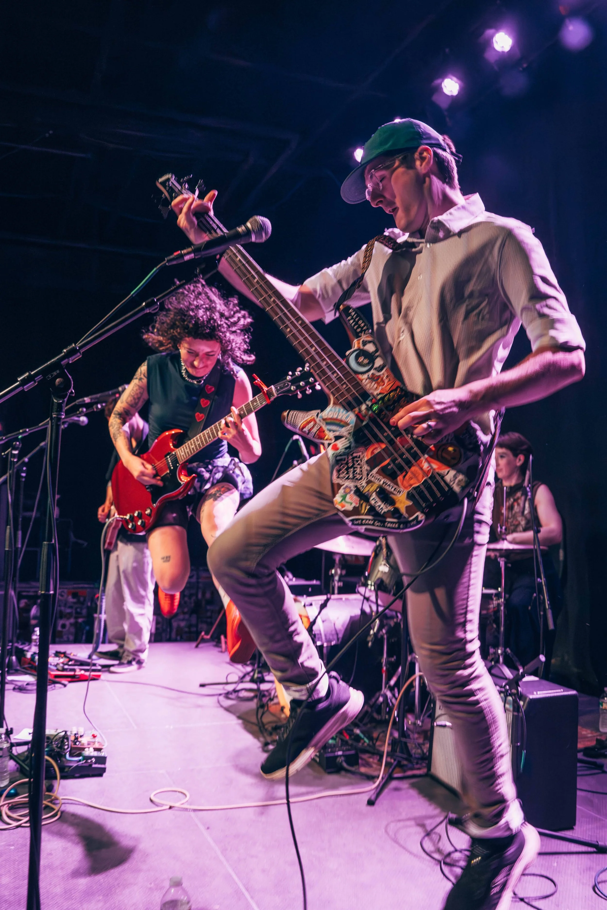 Musicians performing on stage with guitars and equipment, under purple stage lights, with a vocalist and stage monitors.