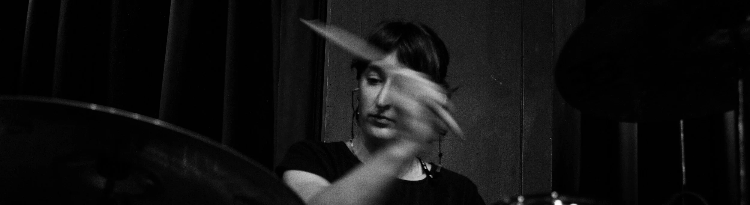 A woman with short dark hair and earrings, playing drums in a dark room.