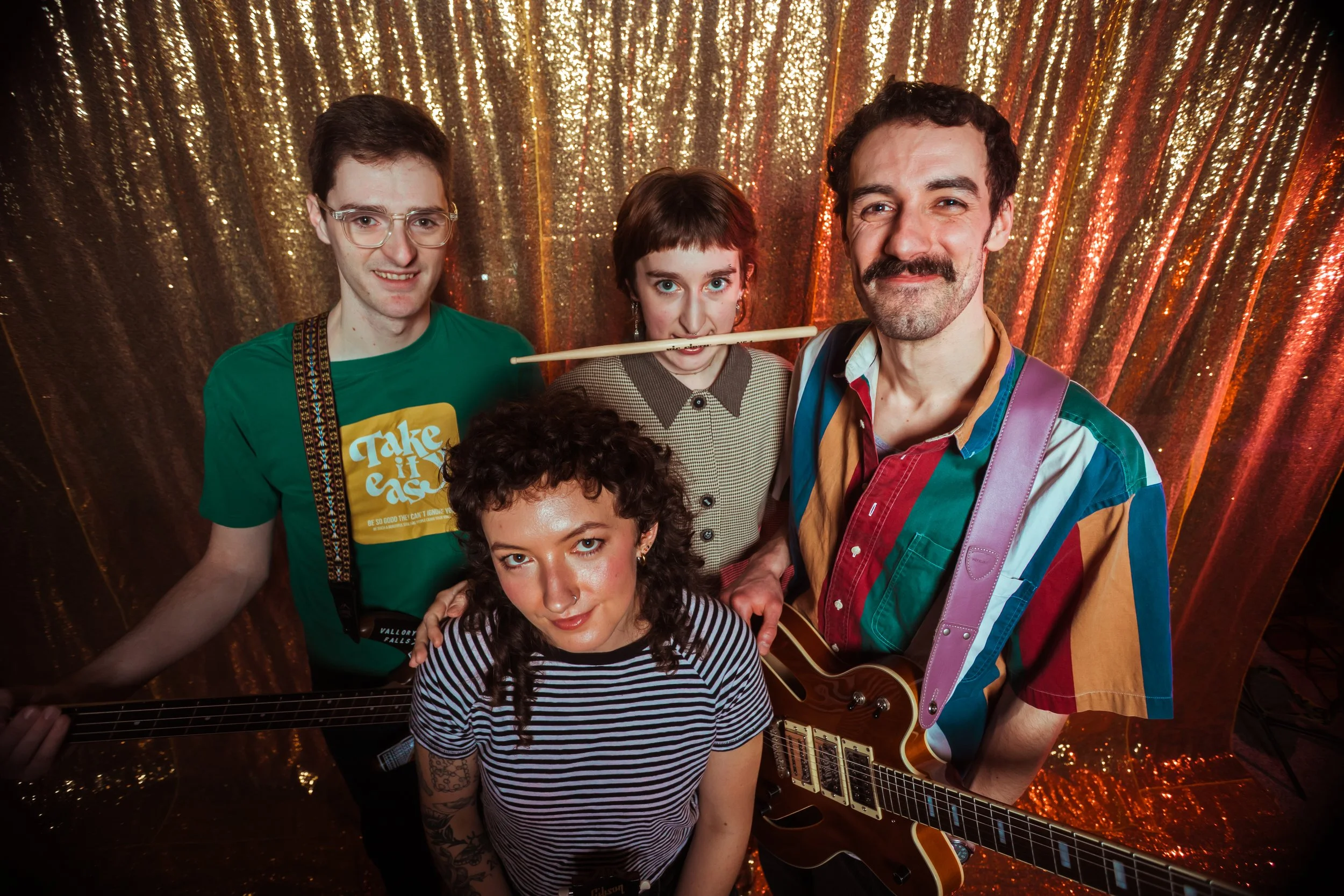Group of five people posing in front of a gold sparkly backdrop, with one person holding a guitar and another wearing drumsticks in their mouth.