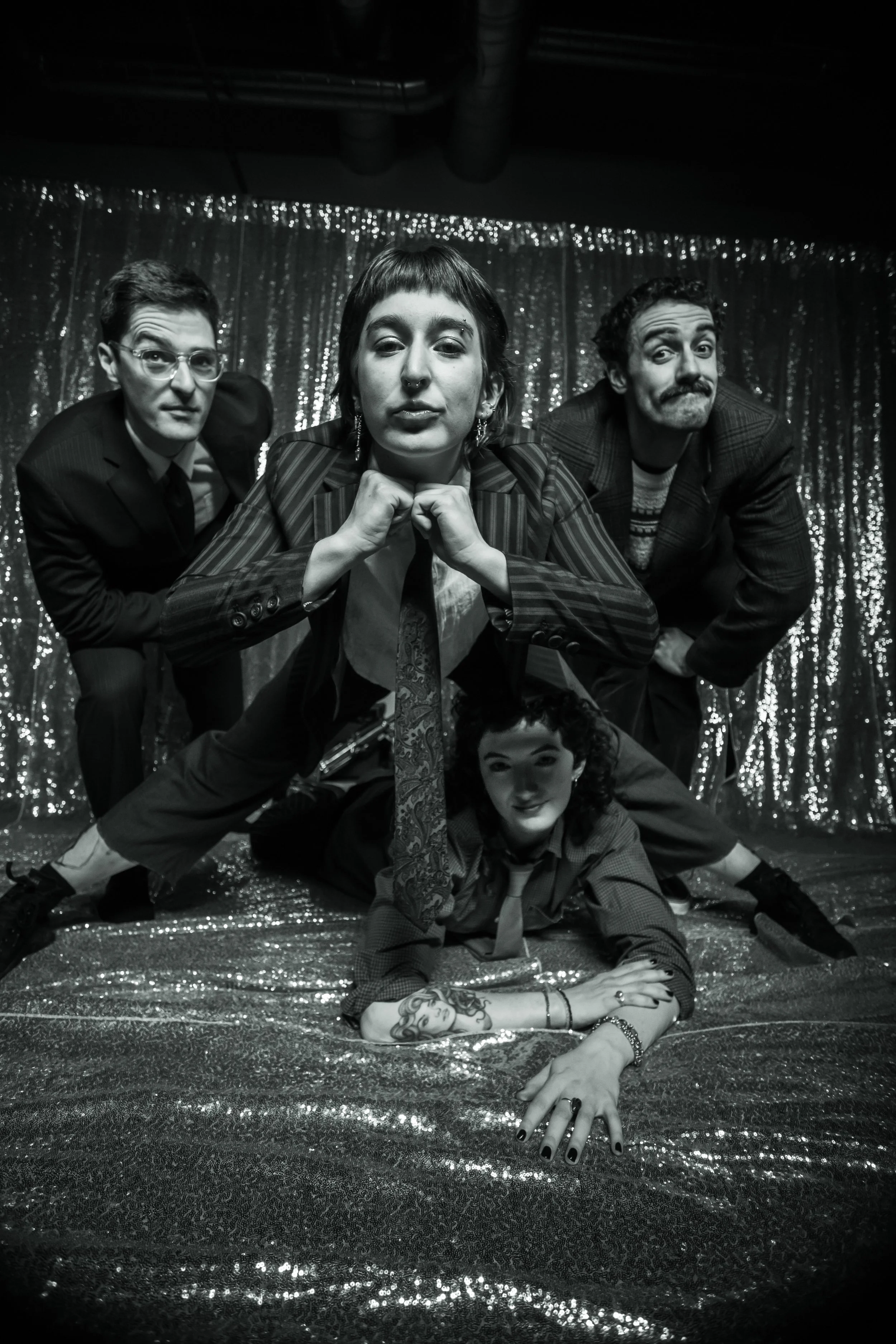 A black and white photo of five young adults posing on a glittery, shiny backdrop with some fabric draped in the background. The group includes two men and three women, with various expressions and poses.
