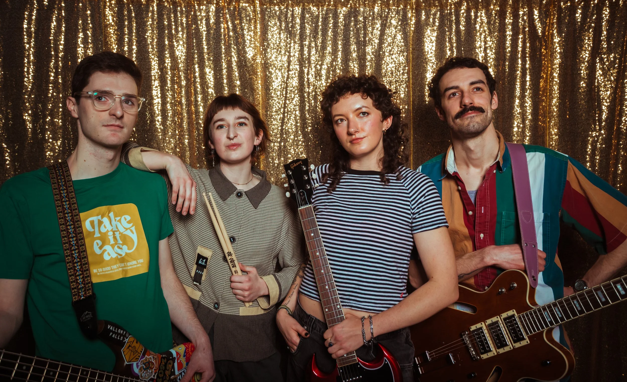 A group of four young adults standing in front of a gold, glittery backdrop. They are holding musical instruments, including guitars and drumsticks, and are dressed casually.
