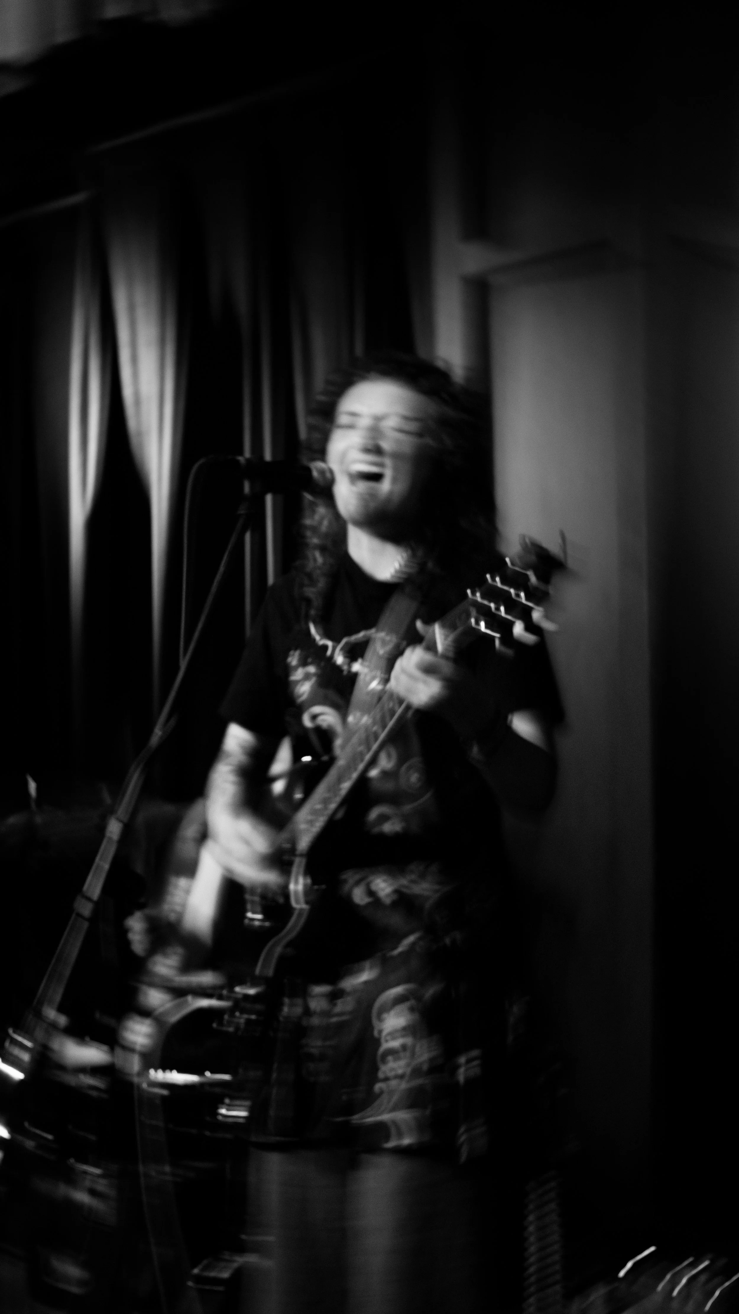 A person singing and playing an acoustic guitar on stage, with a dark background and curtains.