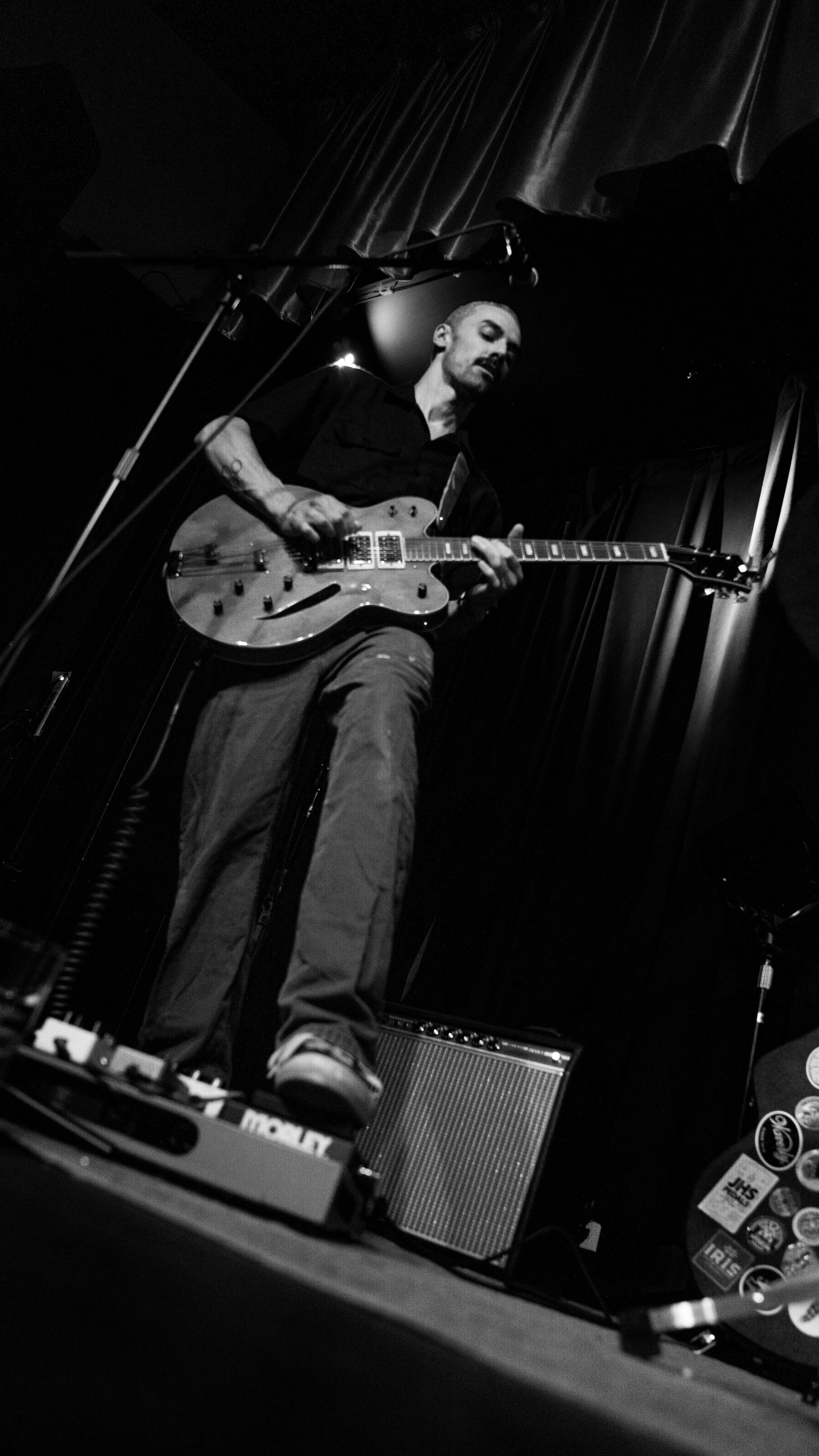 A musician playing an electric guitar on stage, black and white photo, surrounded by musical equipment and dark curtains.