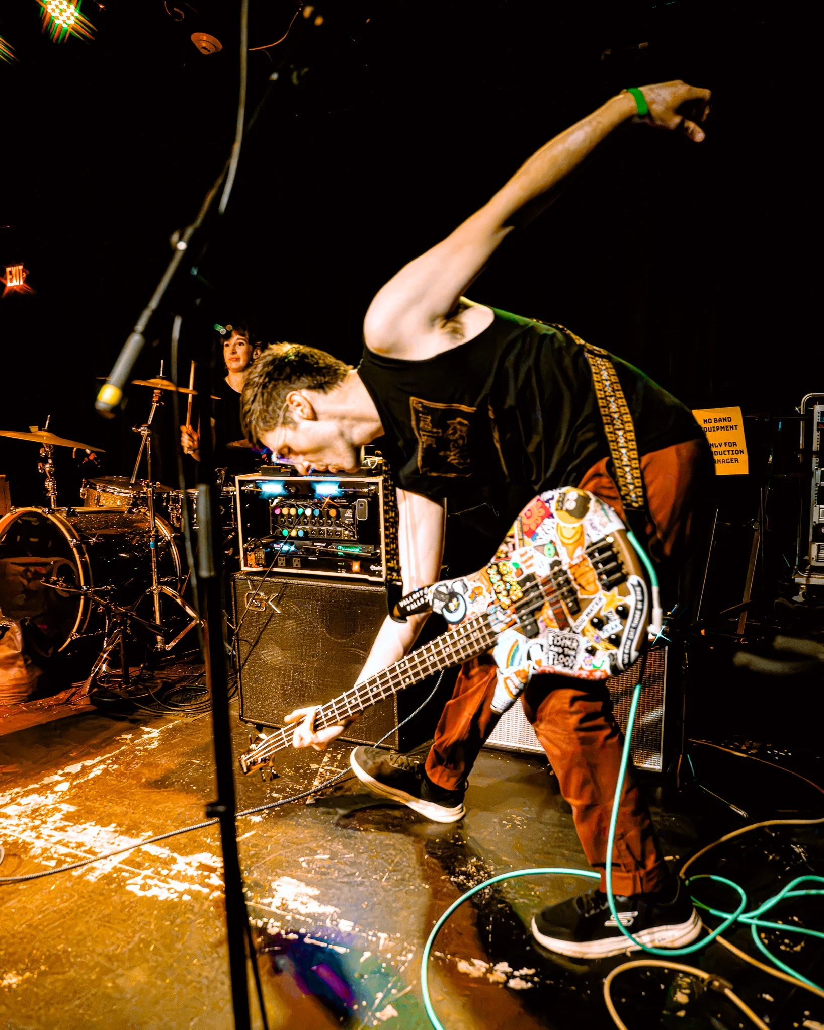 A musician playing an electric bass guitar on a wet stage at night, with a drummer in the background and various musical equipment around, lit by stage lights.