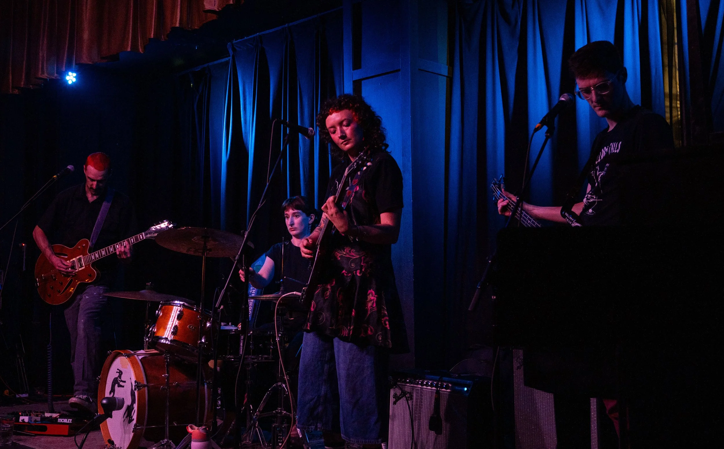 A band performing on stage with blue and red lighting. Three musicians are visible: one with red hair playing an electric guitar, a woman with curly hair and a tattoo playing an acoustic guitar, and a man with glasses and a bass guitar. They are stan