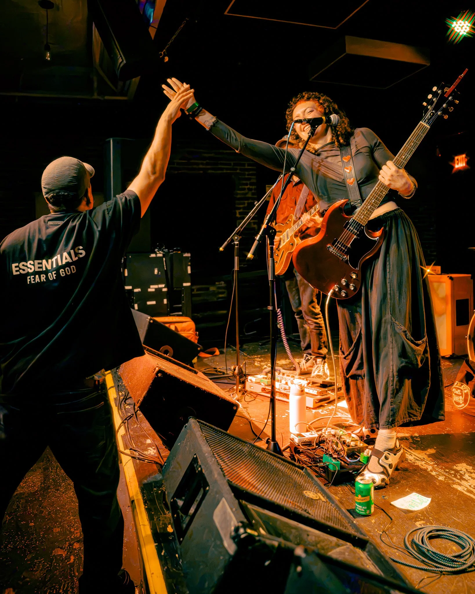 A woman singing into a microphone and playing an electric guitar on stage. She is high-fiving a person in the audience. The stage has musical equipment and the atmosphere is lively with warm lighting.