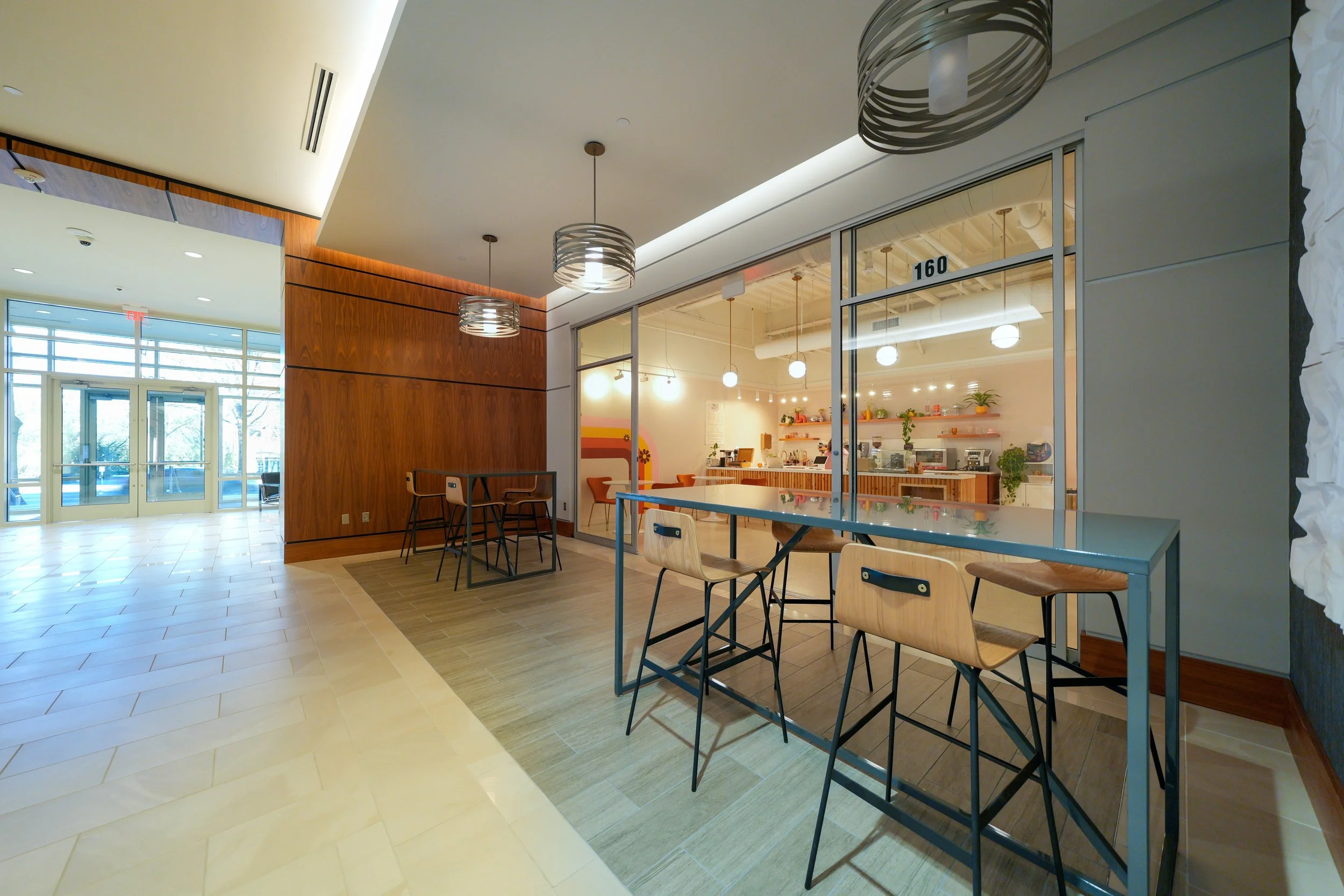 Interior of a modern cafe or dining area with wooden and white walls, bright lighting, and a large window entry door, with counter and seating area visible through glass.