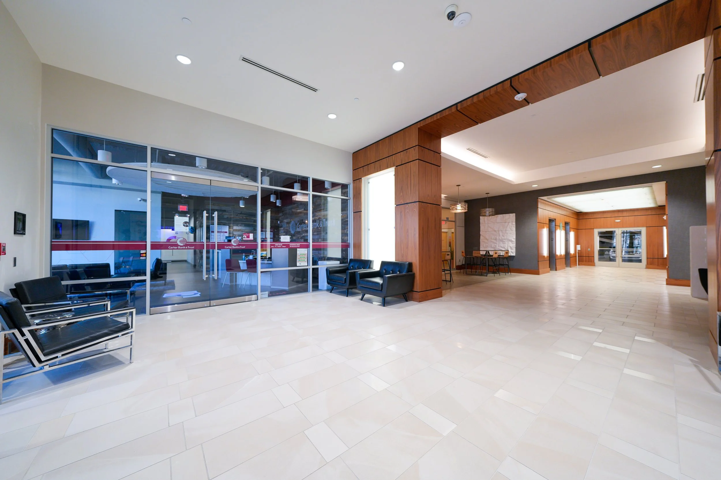 Interior of a modern bank lobby with seating area, tiled floor, wood paneling, glass doors, and a reception area in the back.