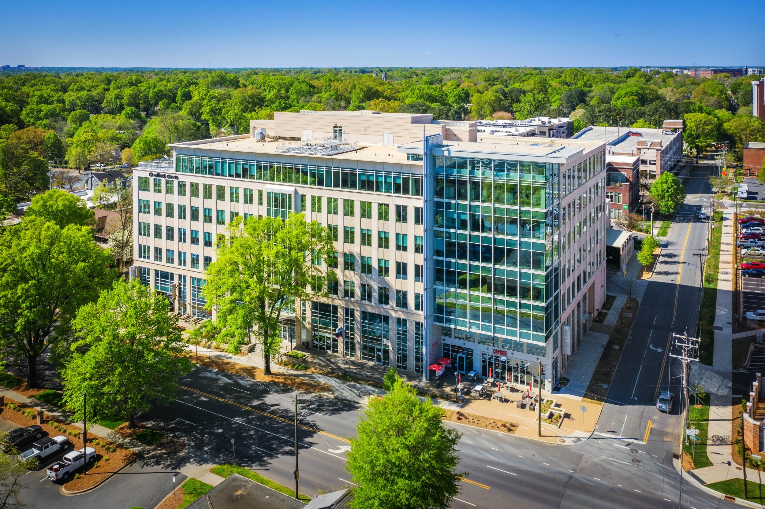 Aerial view of a modern office building with glass windows, surrounded by green trees and a cityscape in the background on a clear, sunny day.