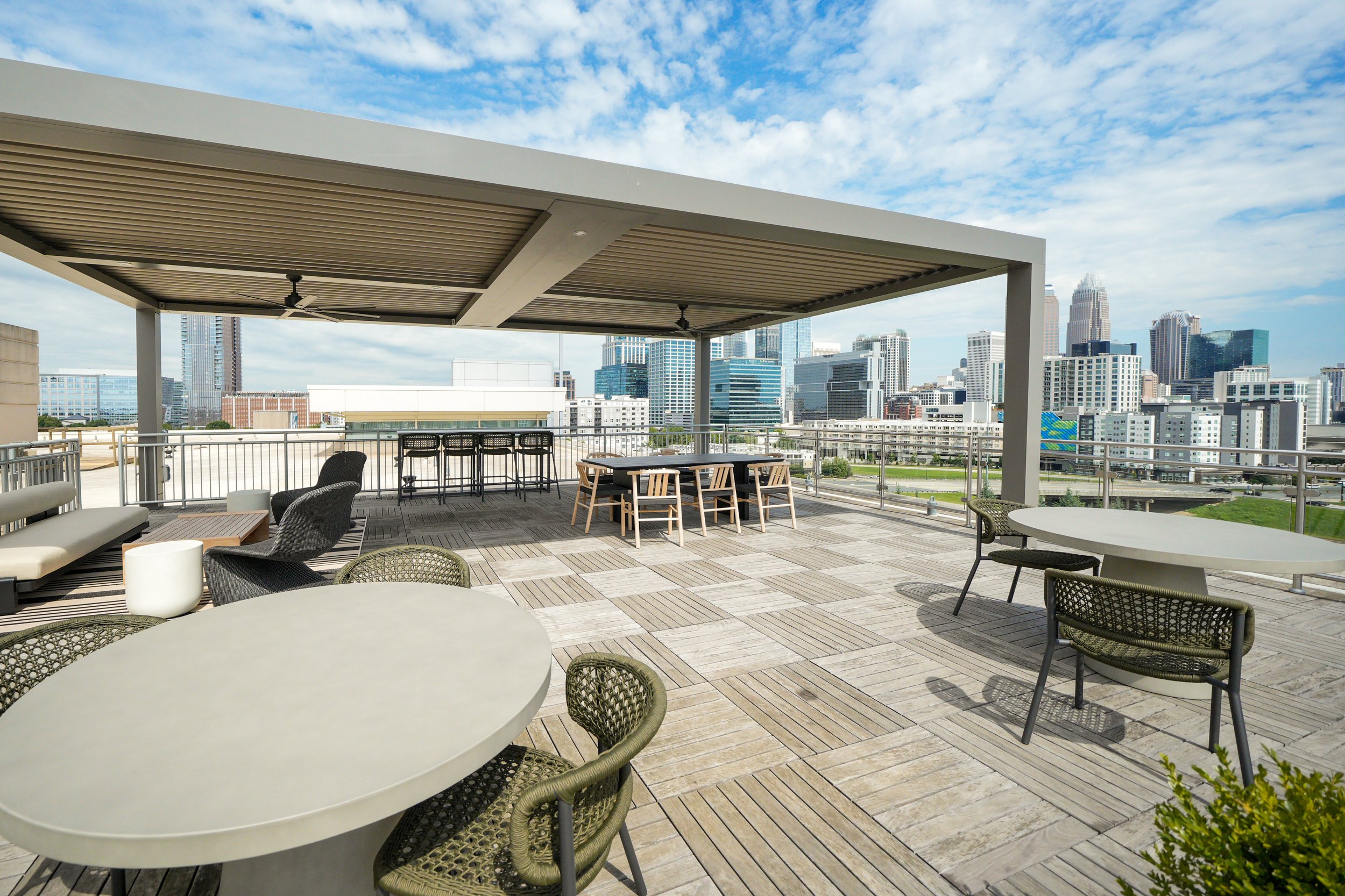 Rooftop patio with outdoor dining and seating area, city skyline with tall buildings in the background, partly cloudy sky.
