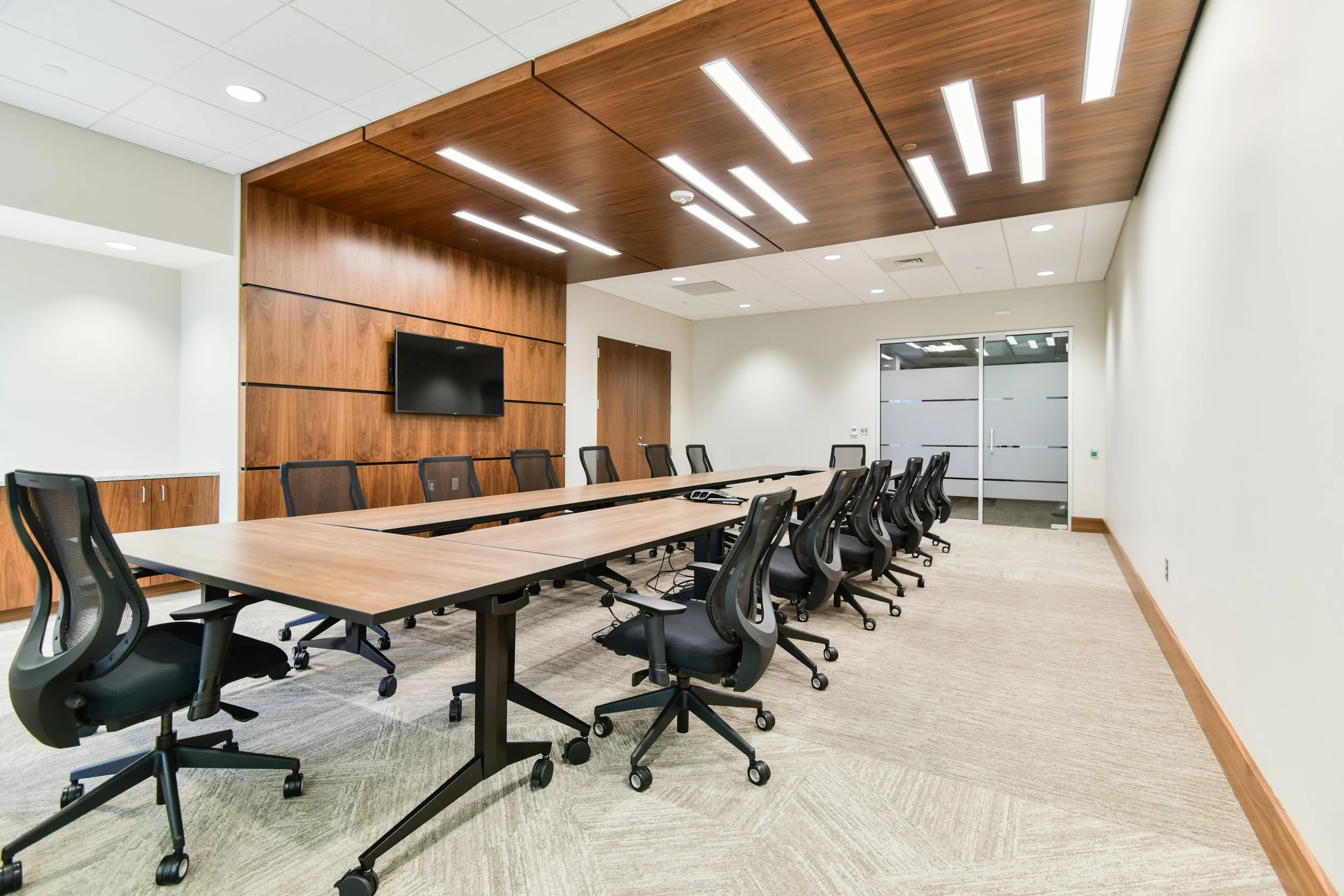 Empty conference room with a long rectangular table surrounded by black office chairs and a wall-mounted TV, with wooden ceiling panels and light-colored walls.