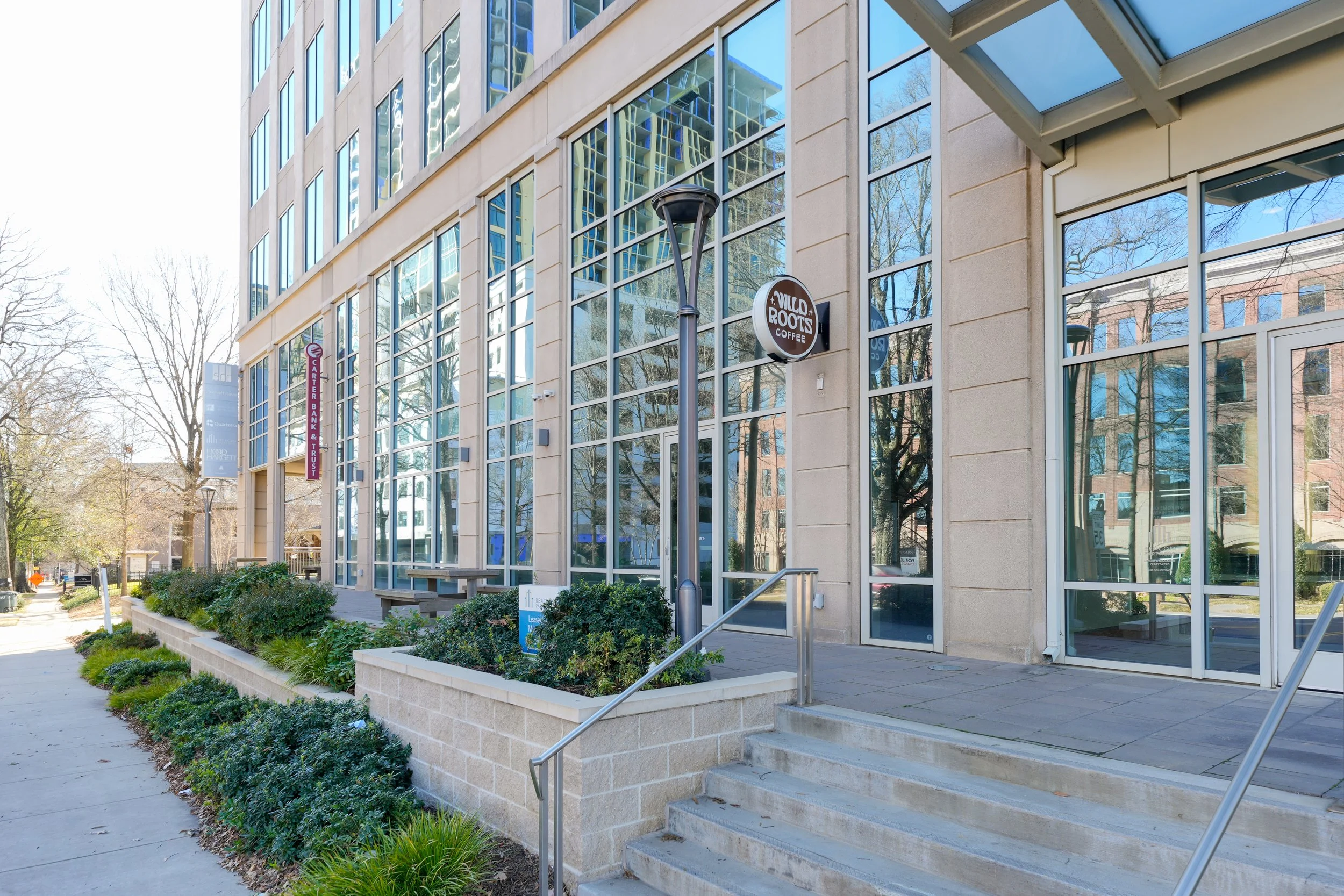 Exterior of a modern office building with large glass windows and a coffee shop sign that says 'Wilco Roots Coffee'. There are steps and a ramp leading to the entrance, along with landscaped bushes and trees nearby.
