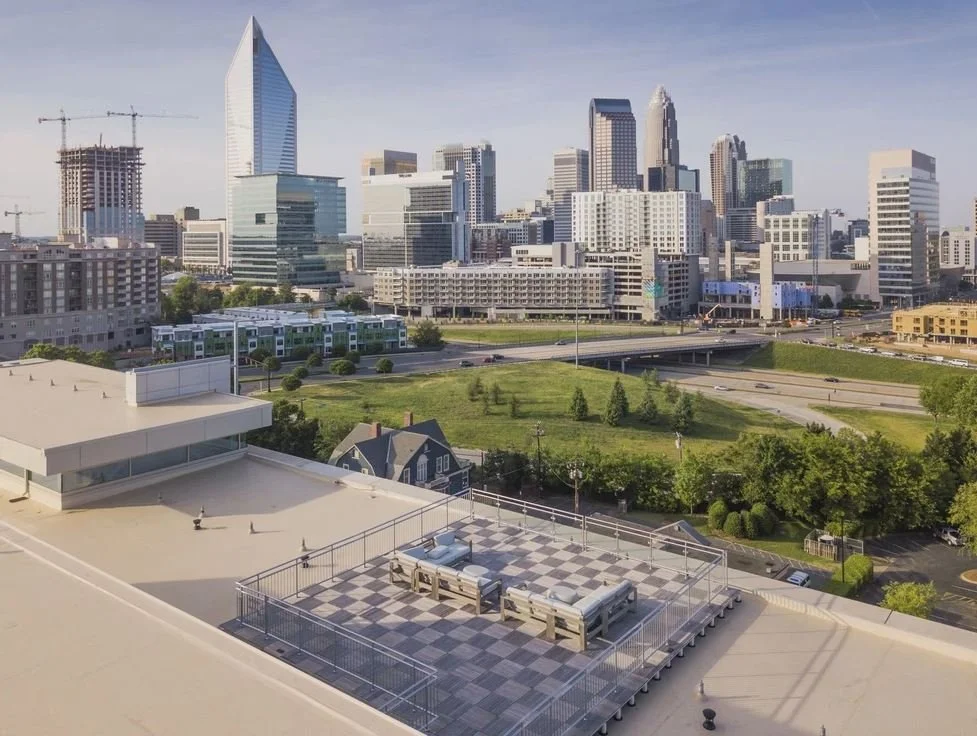 City skyline featuring tall modern skyscrapers, some under construction, with a rooftop area in the foreground that has seating and a checkered floor, and a grassy park with trees below.