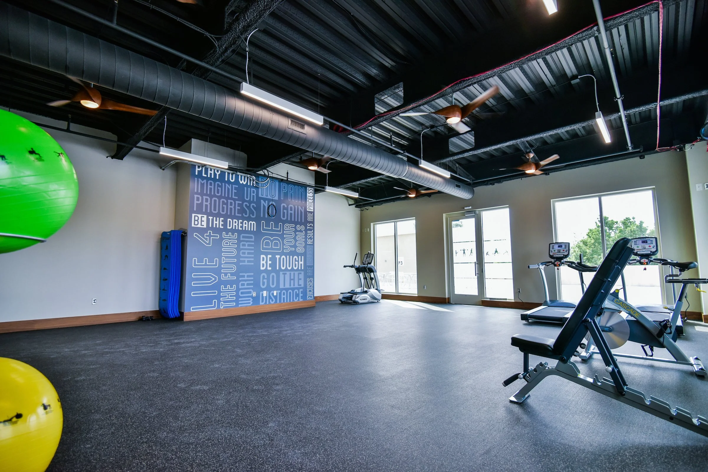 A gym interior with exercise machines, large windows, motivational wall art, and fitness equipment including a green exercise ball, yellow ball, and treadmills under a black ceiling with exposed ductwork and ceiling fans.