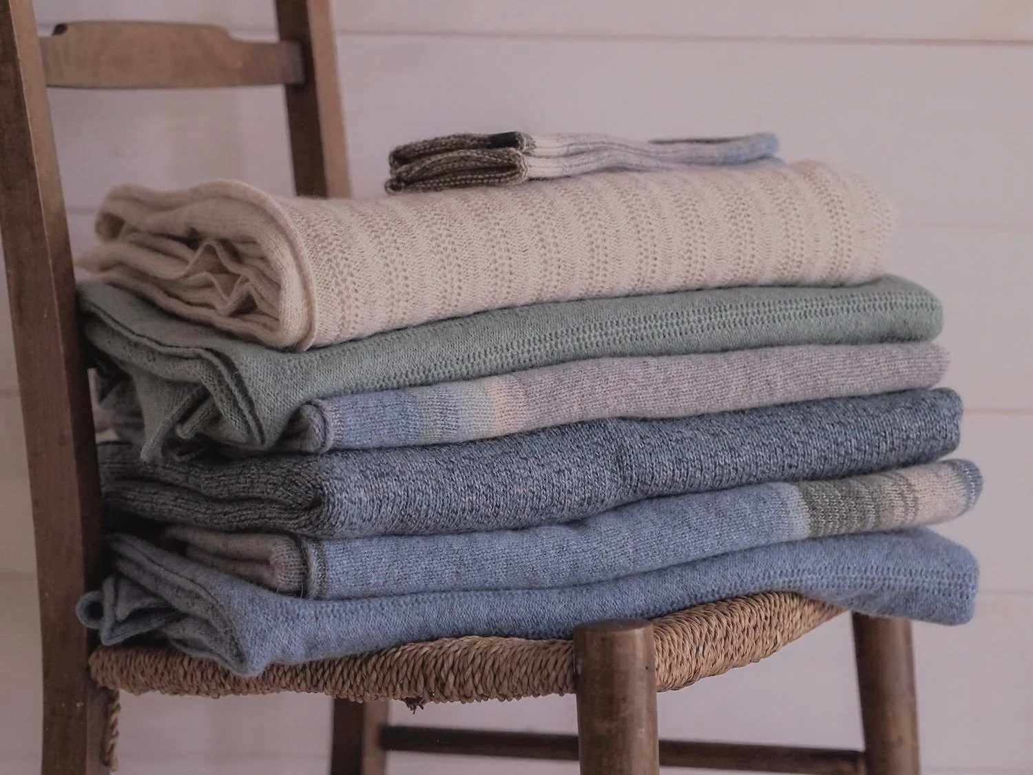 Stack of neatly folded knitted, woven, and fabric towels or blankets in neutral colors on a wooden chair.